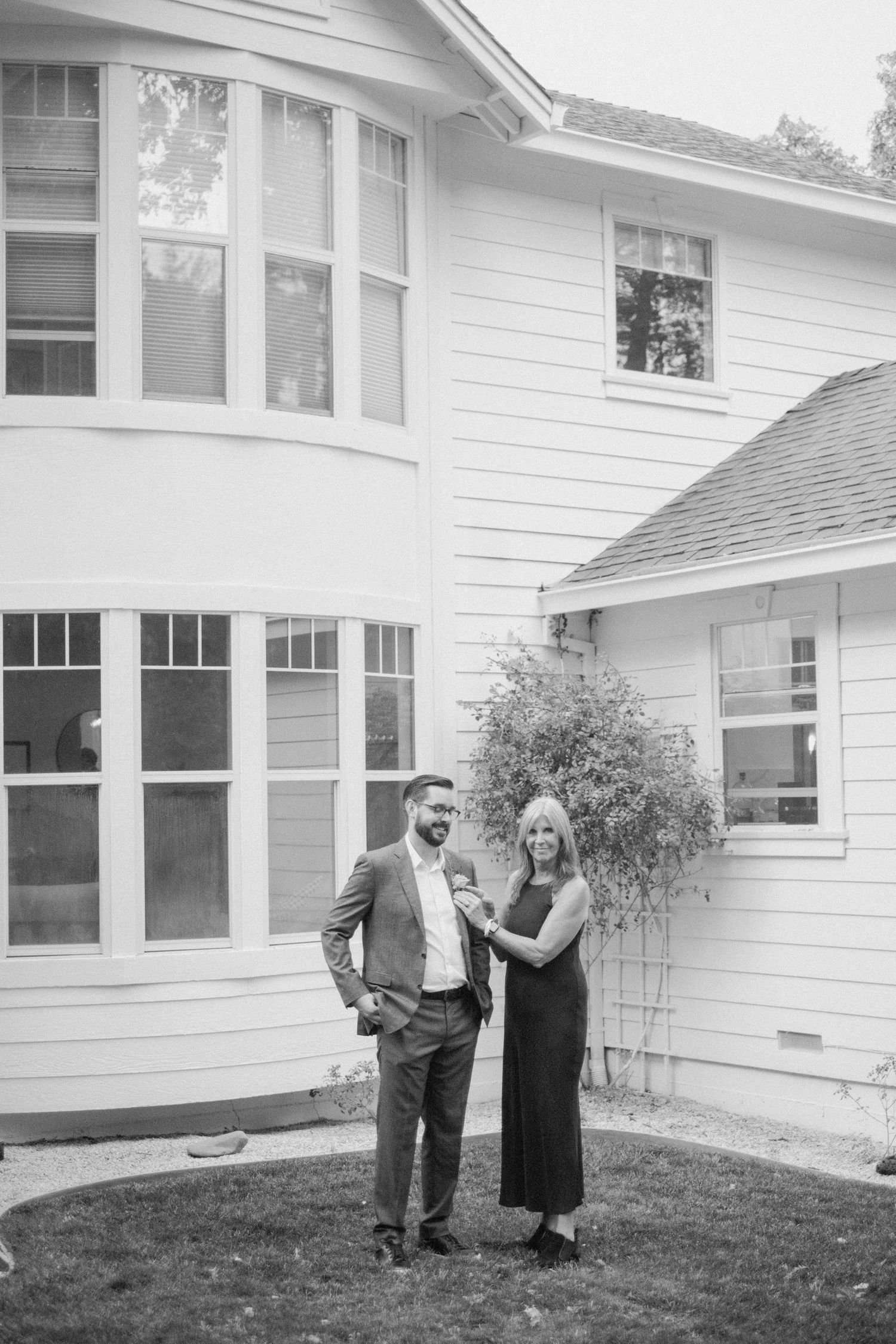 Couple posing together in black and white photo outside elegant two-story white house with bay windows.