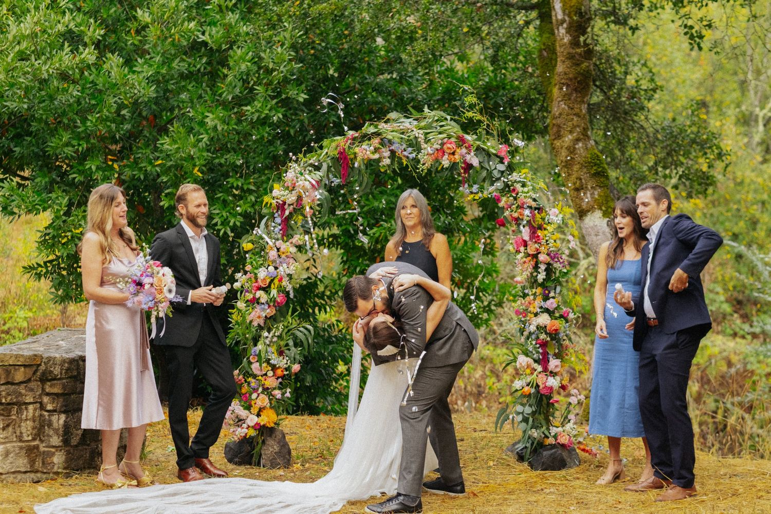 Wedding couple sharing first kiss under colorful floral arch surrounded by guests outdoors.