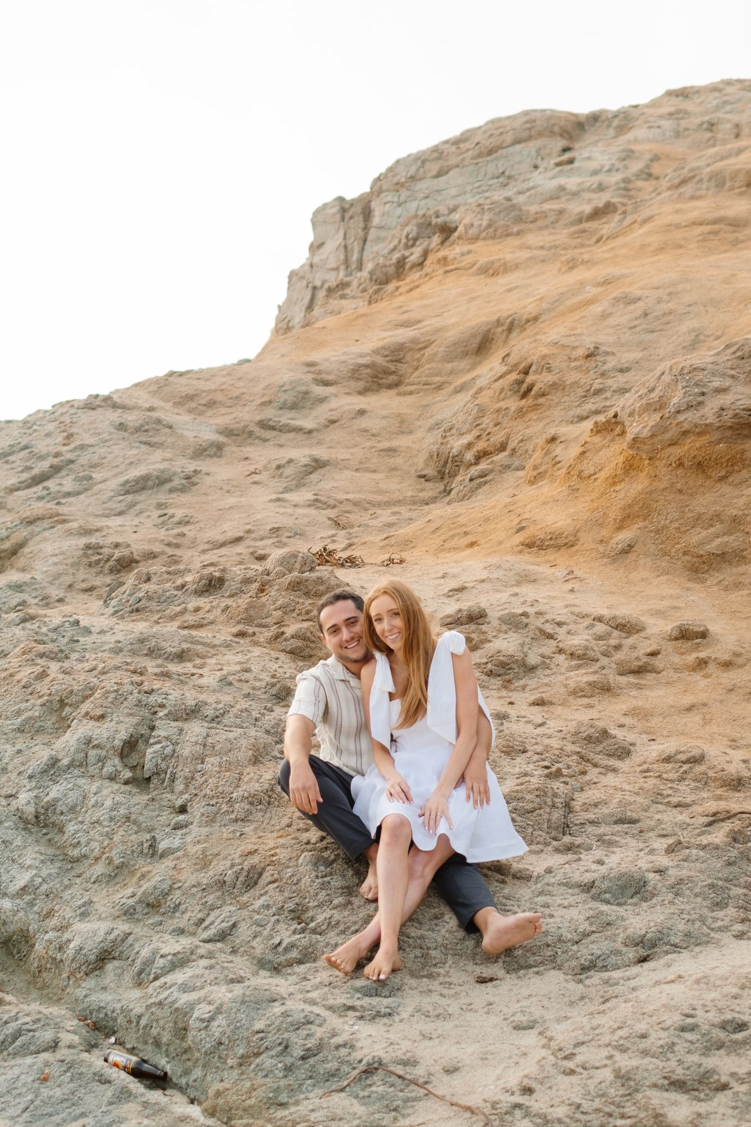 Couple sitting together barefoot on sandy rocky cliffside, woman in white dress leaning on man.