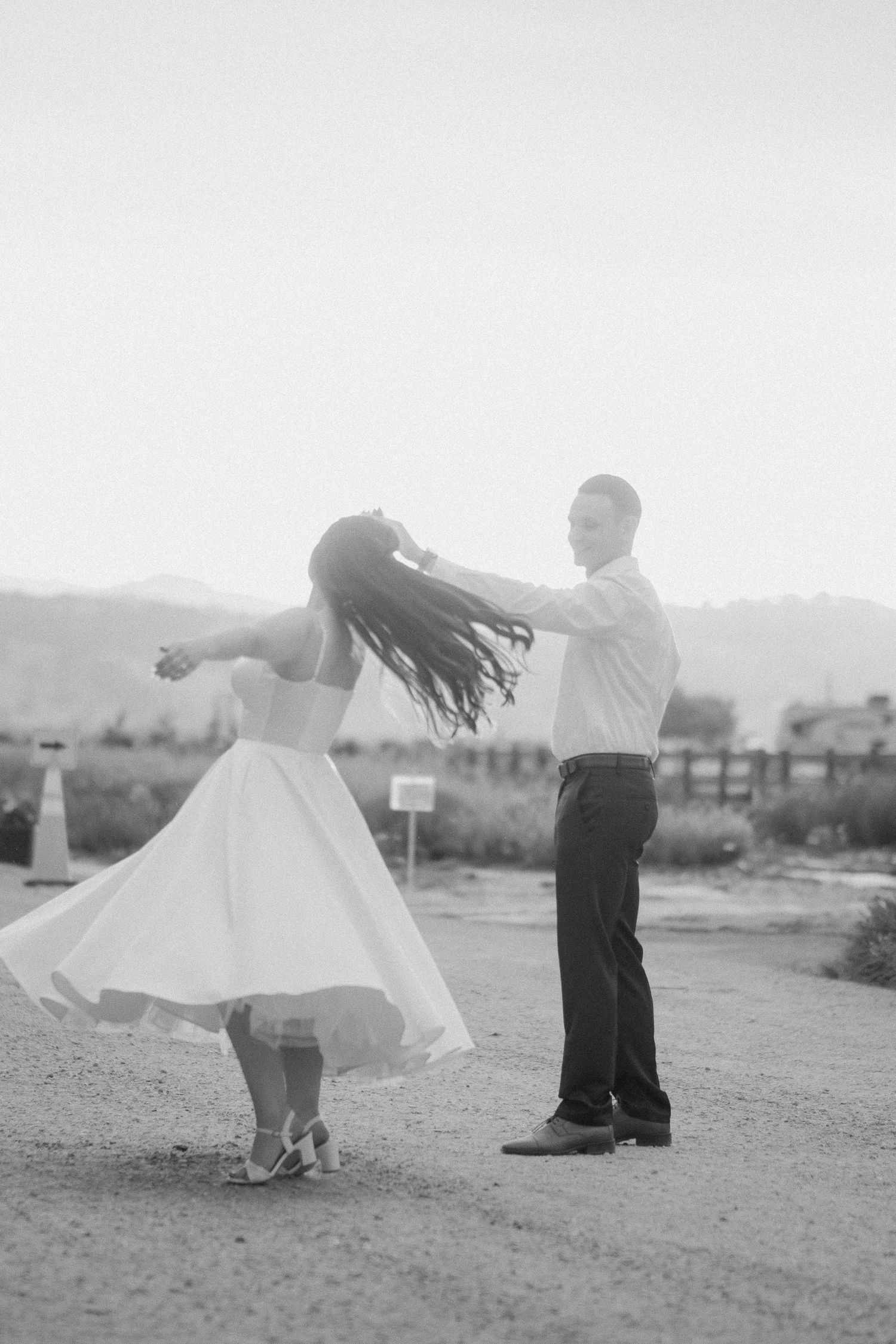 Black and white photo of groom twirling bride on a dirt path, her dress and hair flowing in the breeze.