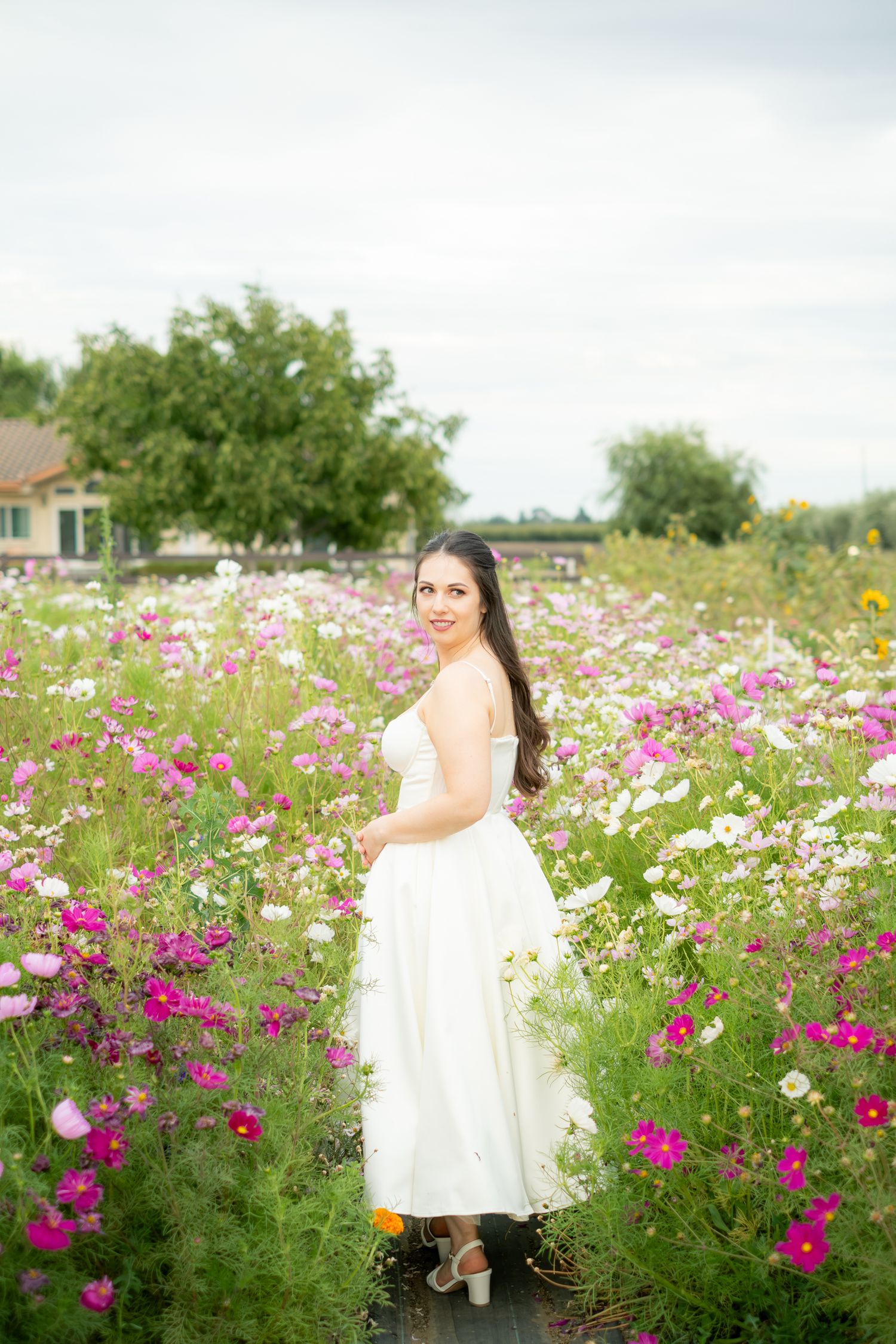 Young woman in white dress smiling in a vibrant cosmos flower field.