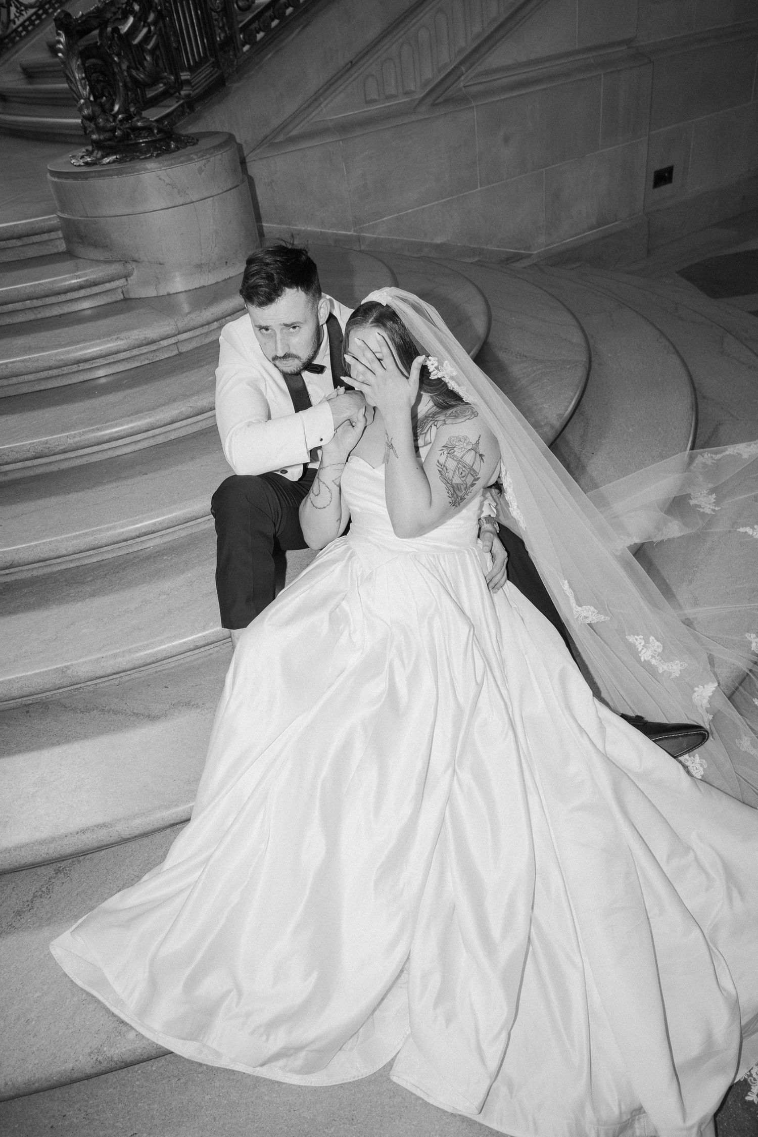 Black and white photo of bride and groom sitting on grand staircase, bride covers her face with hands emotionally.