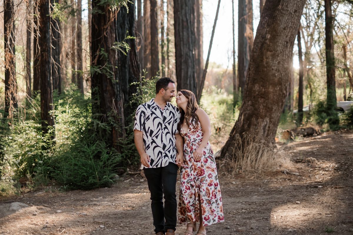 Couple sharing a romantic moment on a forest trail surrounded by tall trees and dappled sunlight.