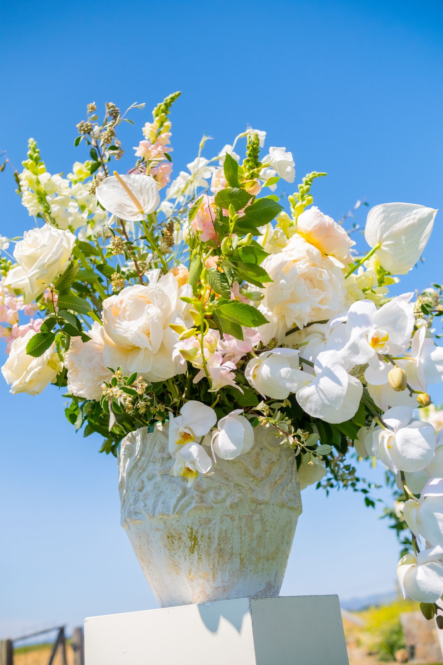 Elegant white and blush floral arrangement in a vintage vase against a clear blue sky.