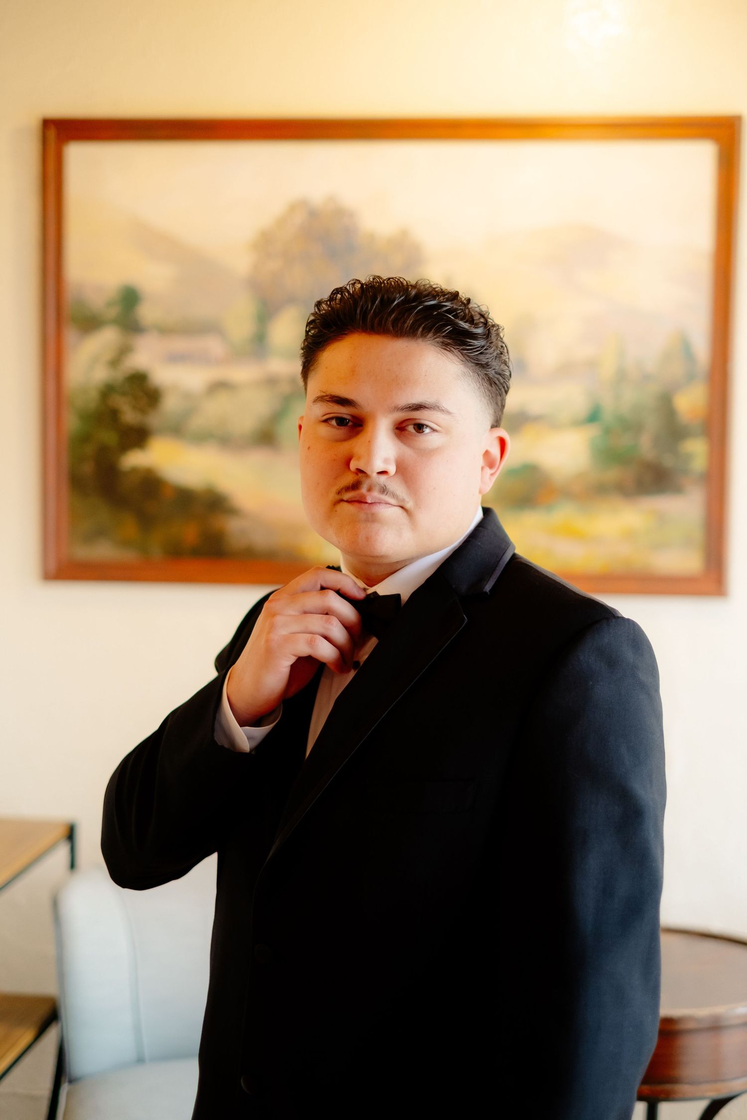 Groom in black tuxedo adjusting bow tie before wedding ceremony with landscape painting in background.