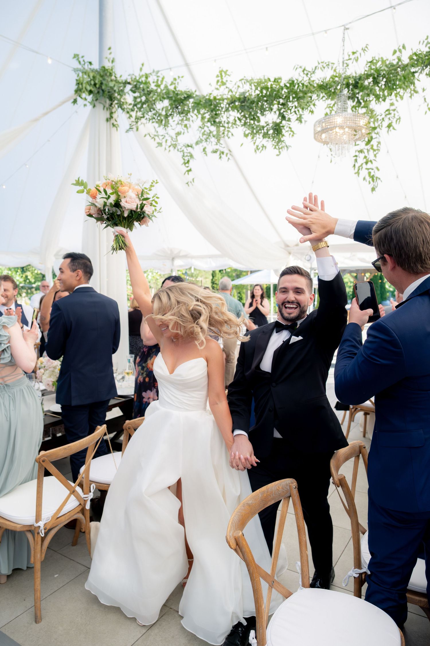 Bride and groom celebrate joyfully entering their tented reception, raising hands and bouquet to cheering guests.