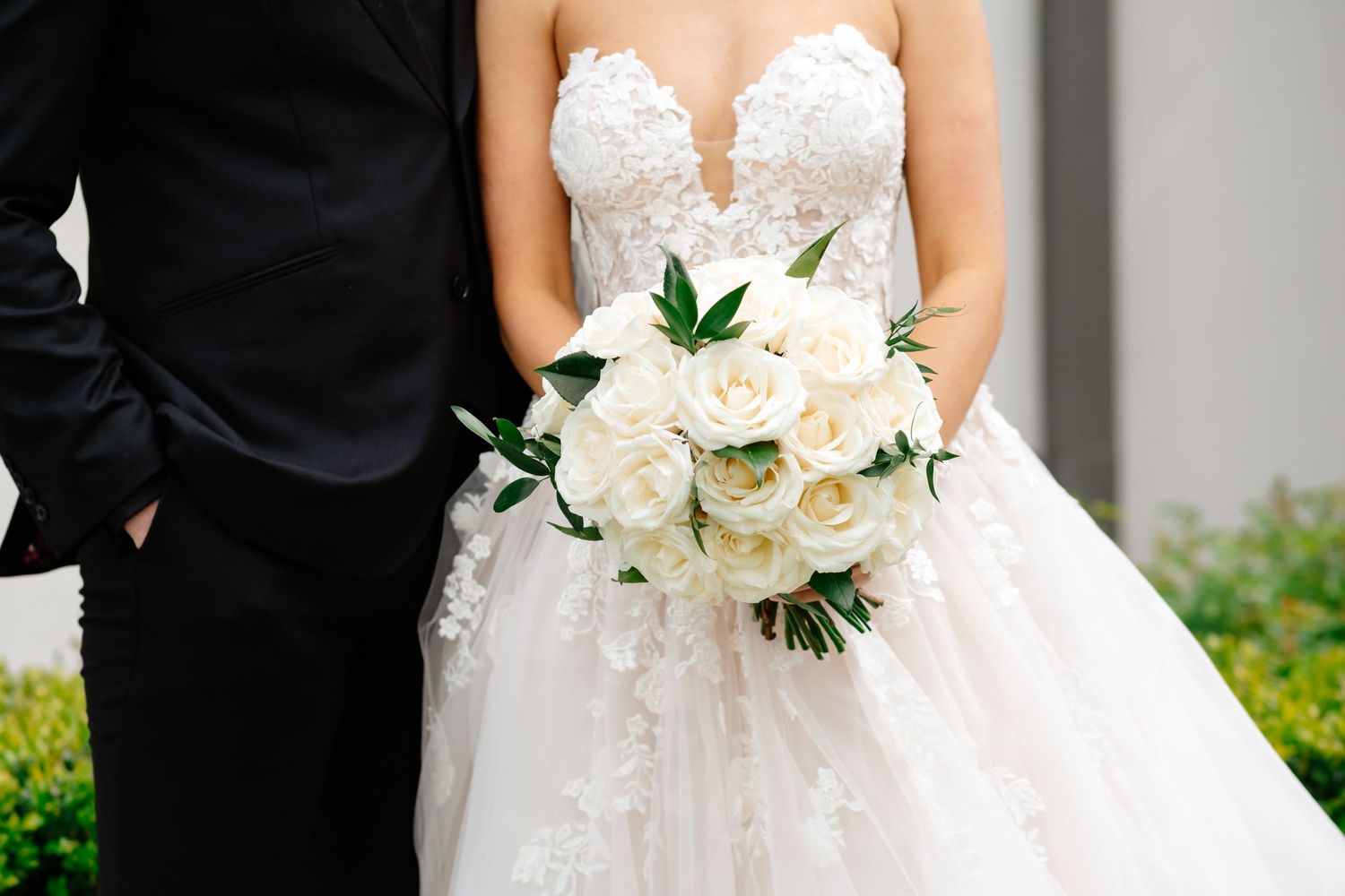 Bride in lace gown holds white rose bouquet beside groom in black tuxedo.
