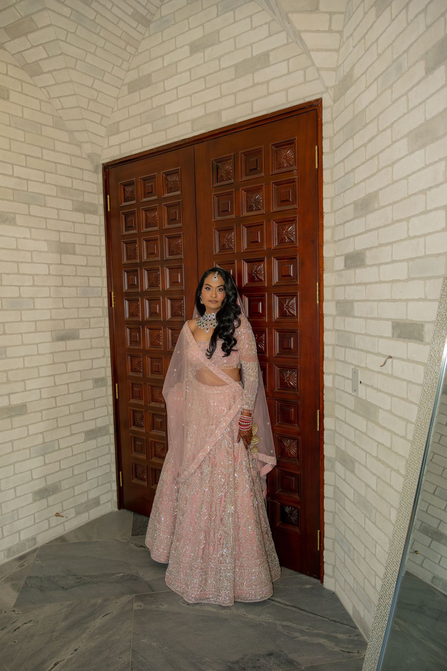 Bride in pink lehenga standing before wooden doors in a marble hallway.