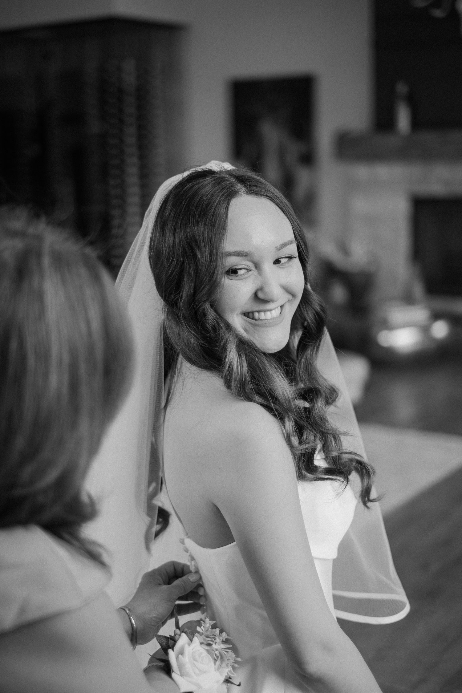 Smiling bride in white gown and veil being helped with her dress in black and white photo.