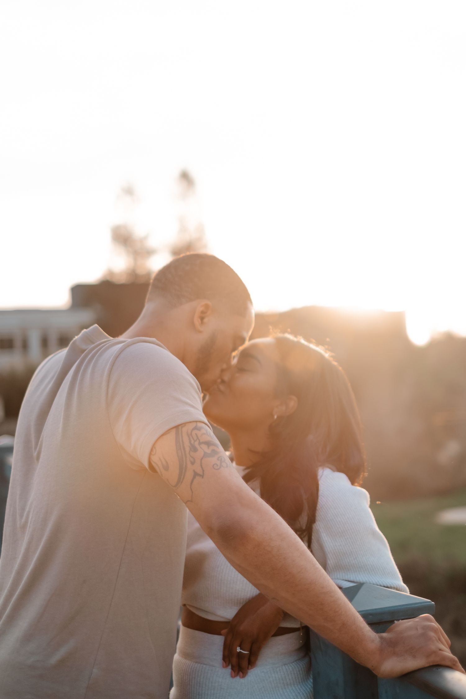 A couple shares a tender kiss at sunset on a bridge, bathed in warm golden backlight.