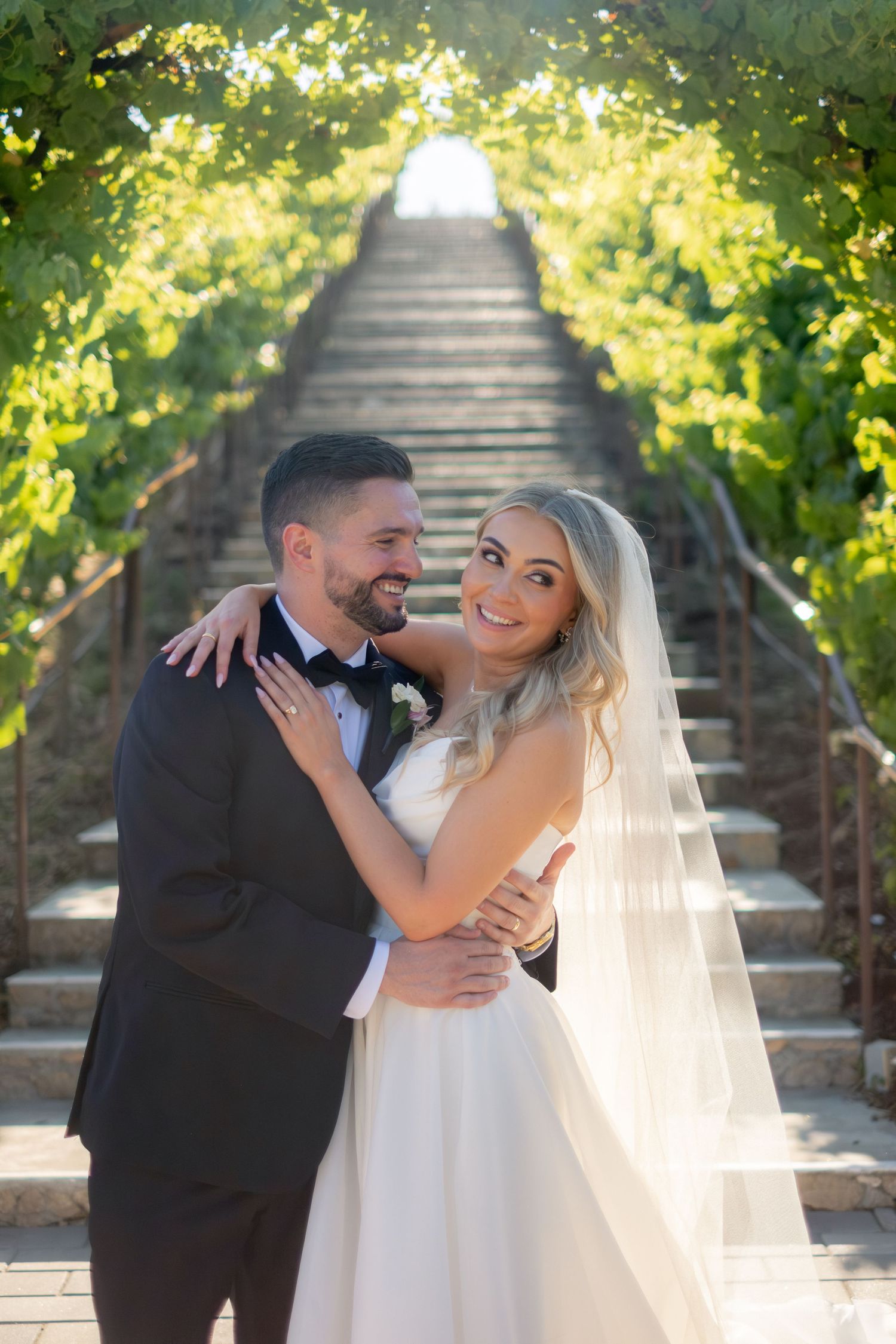 Smiling bride and groom embrace under a lush green vine-covered staircase at vineyard wedding.