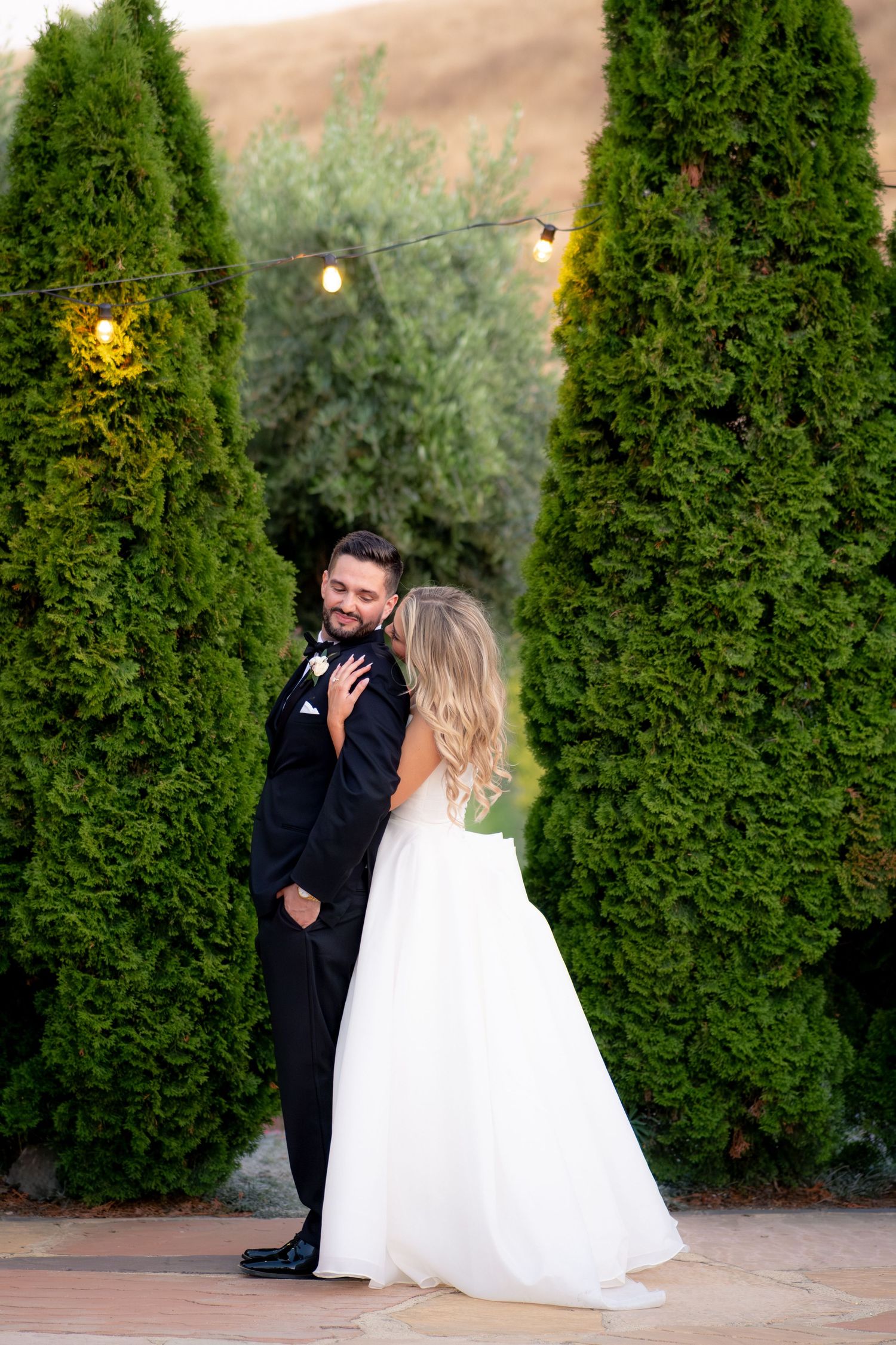 Bride and groom share a romantic embrace among tall green cypress trees with string lights at their outdoor wedding.