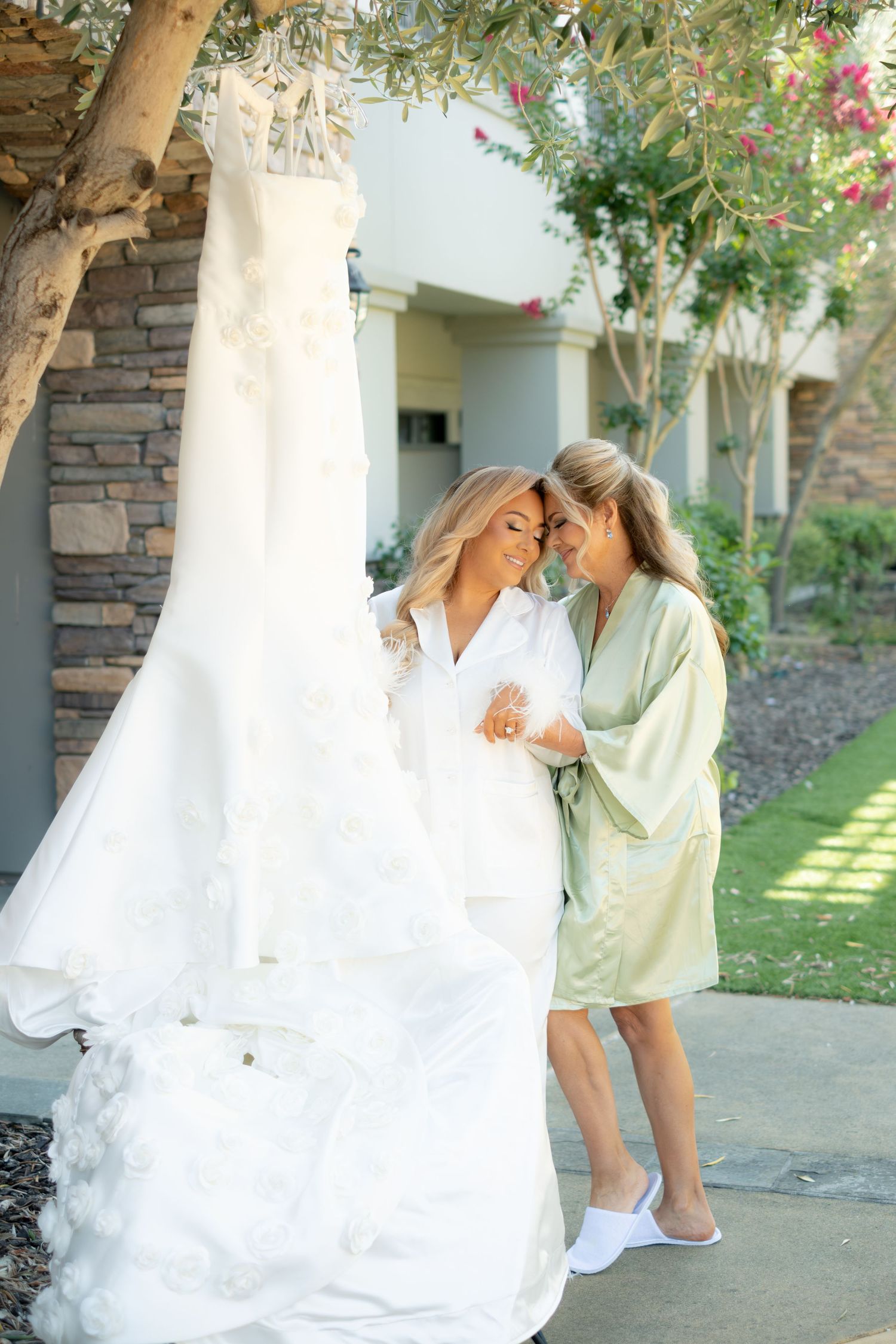 Bride and bridesmaid in robes share an intimate moment beside a hanging floral wedding dress outdoors.