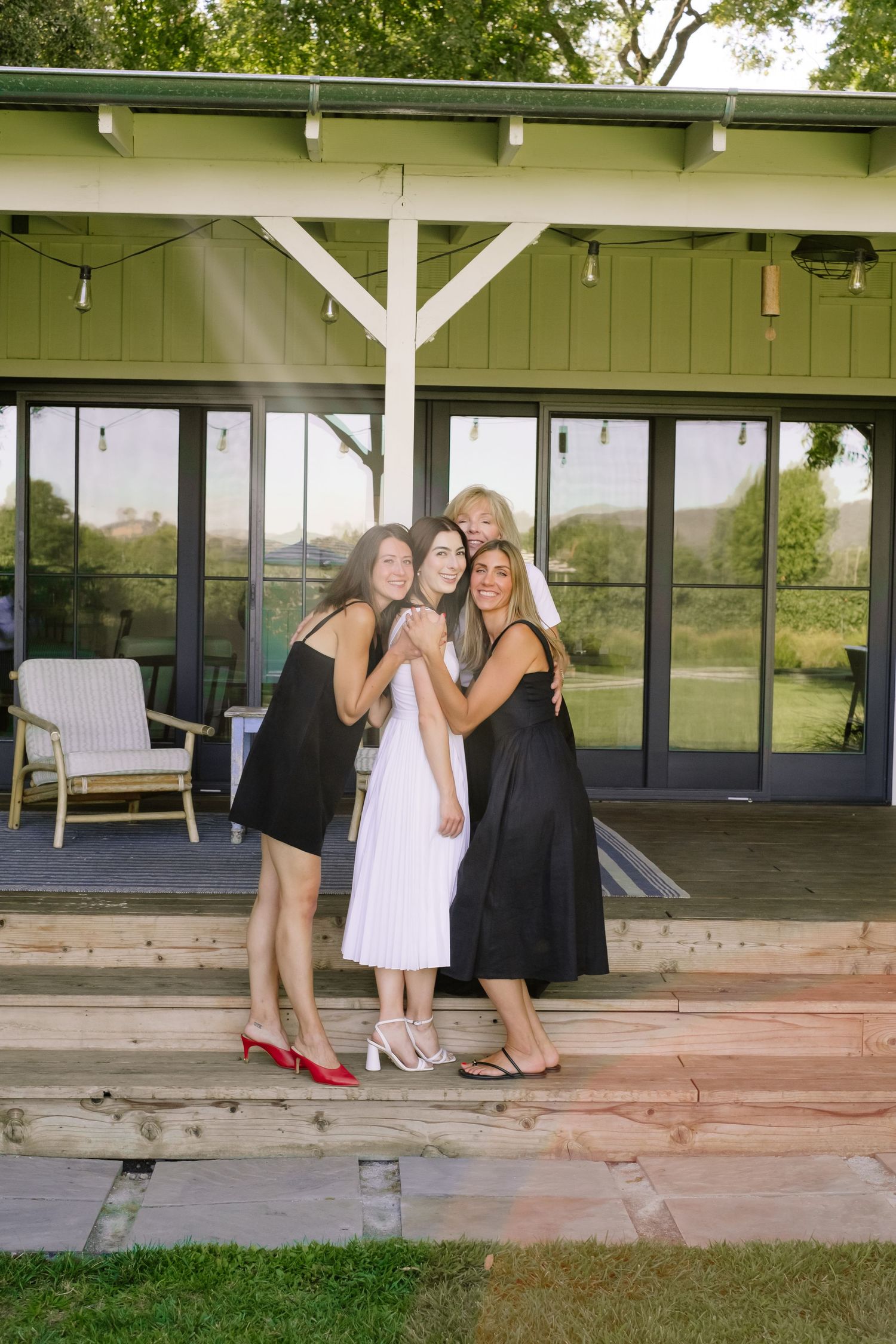 Four women in black and white dresses pose together smiling on wooden deck outside green building.
