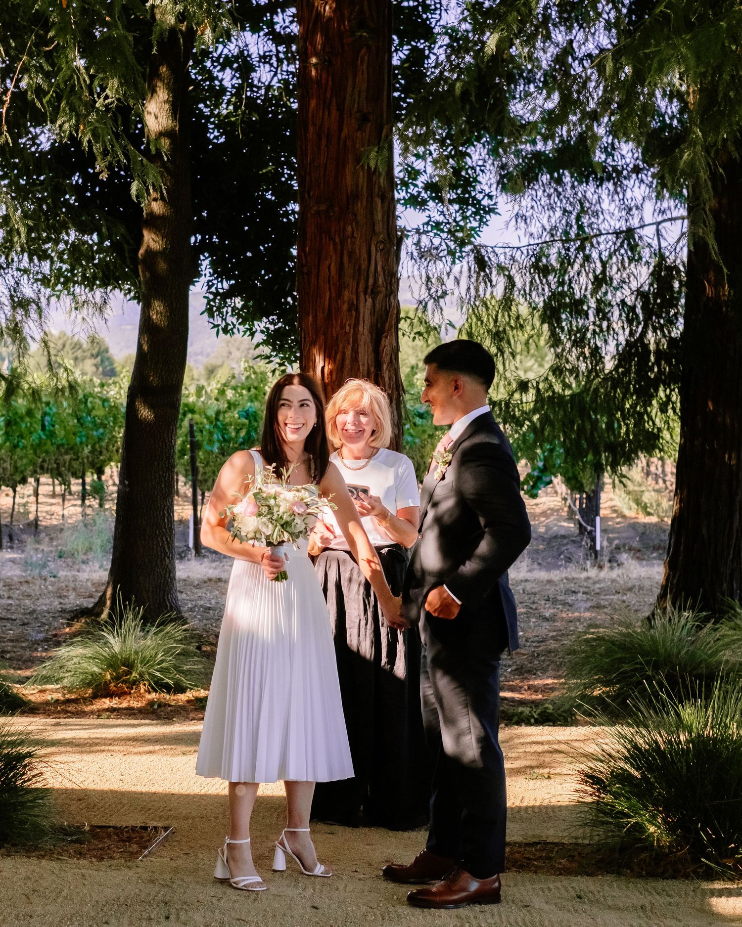Bride in white midi dress and groom in navy suit with officiant during outdoor wedding ceremony.