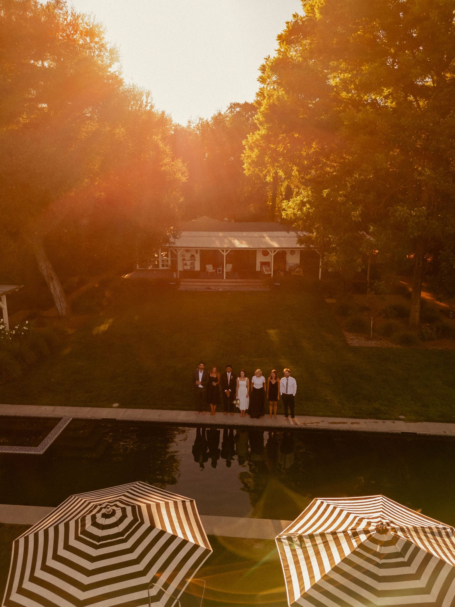 Aerial view of wedding party standing by reflective pool at golden sunset with striped umbrellas.