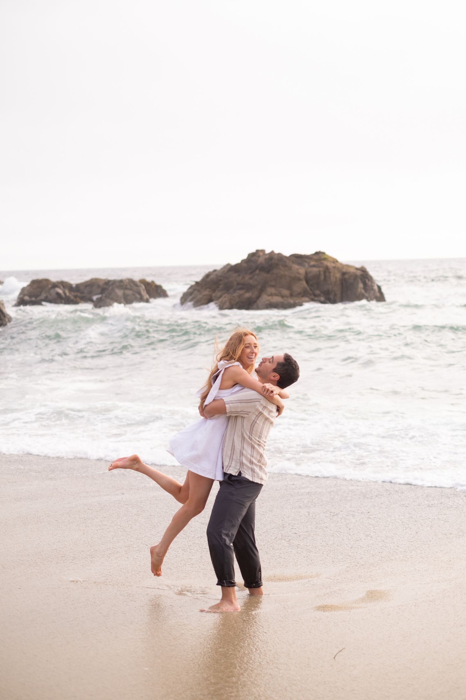 Romantic couple embracing on wet sandy beach with man lifting woman in white dress near ocean waves.