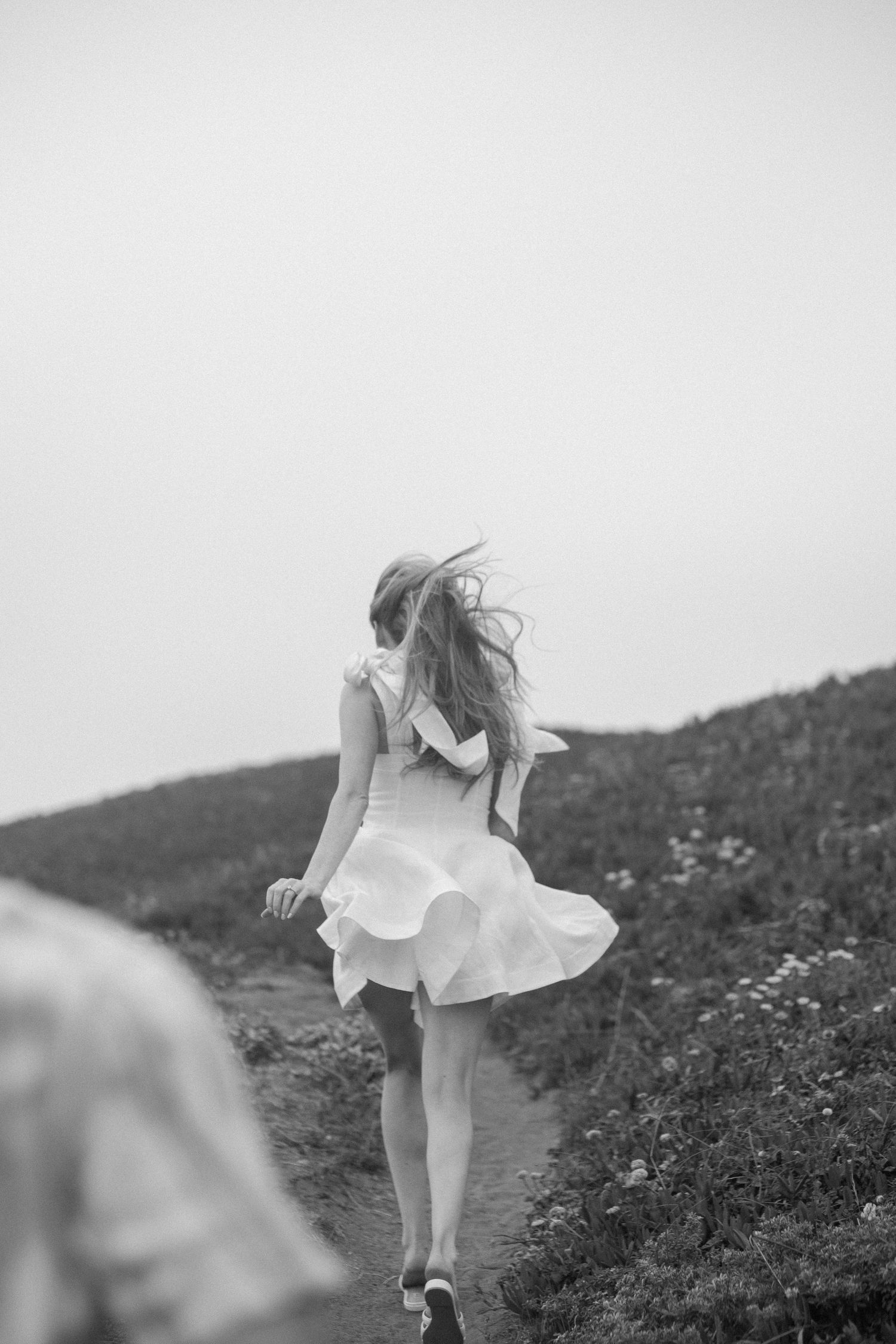 Black and white photo of a woman in a white dress walking up a hillside path, hair blowing in the wind.
