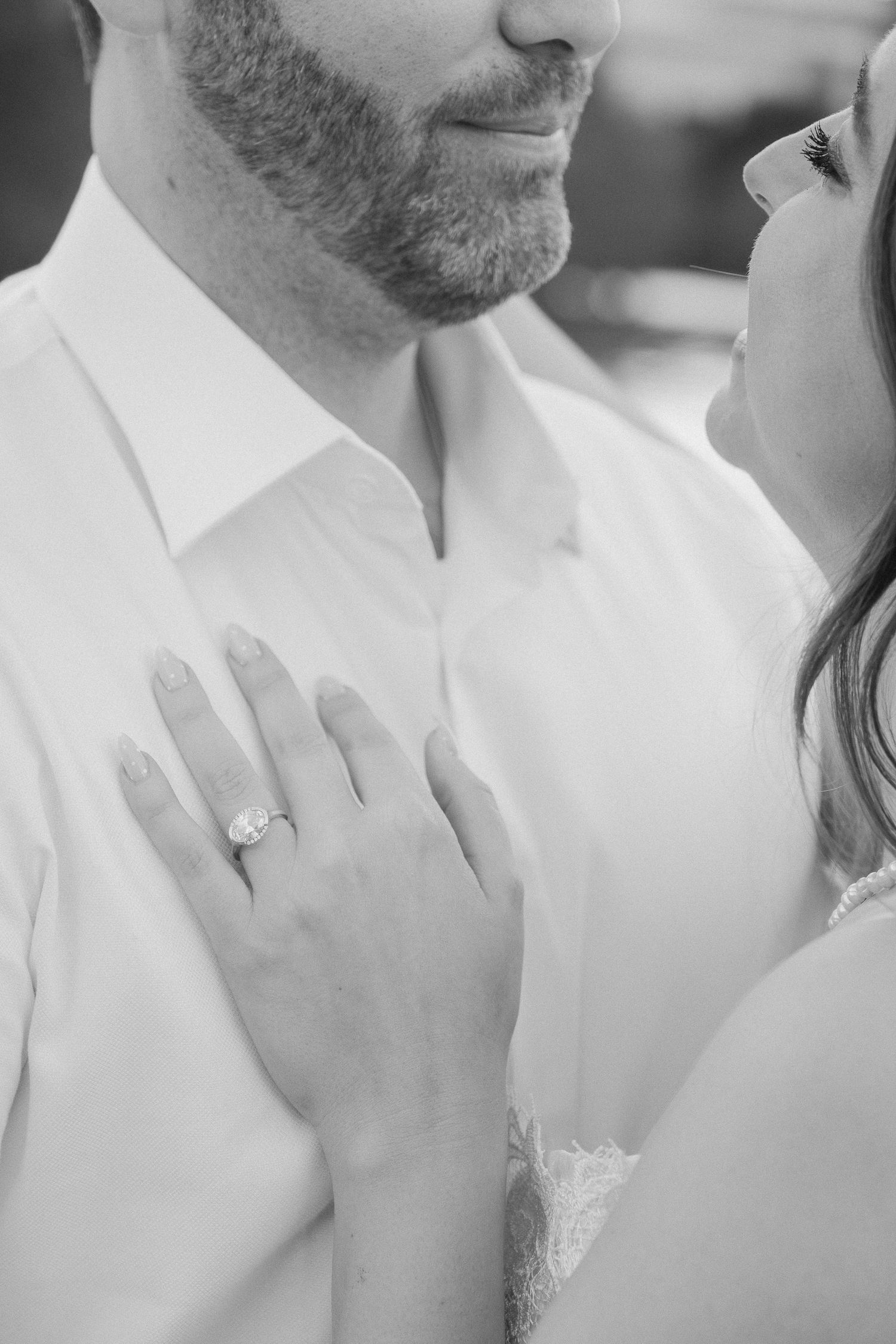 Black and white close-up of an engagement ring as bride rests her hand on grooms chest.