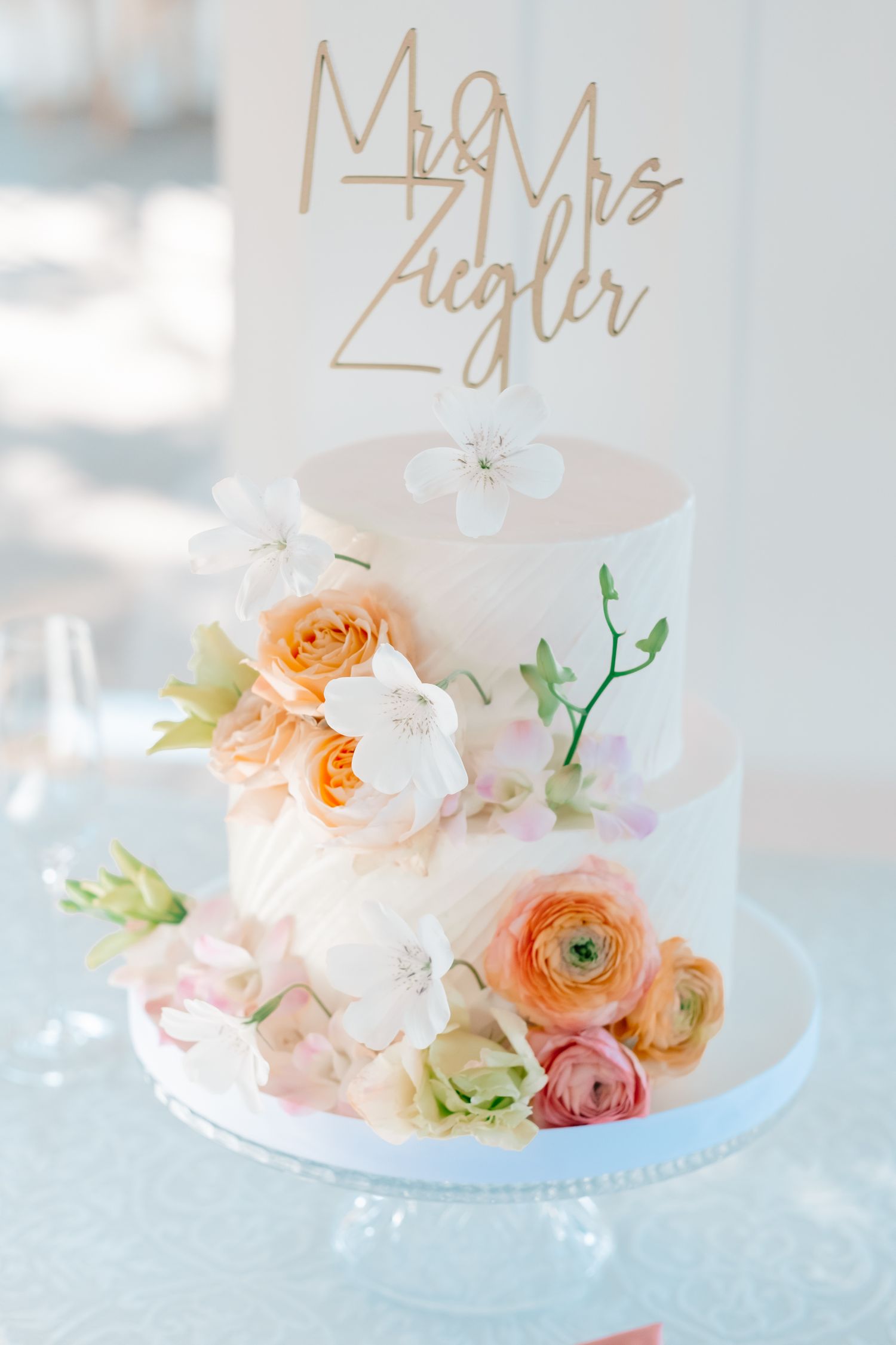 Two-tier white wedding cake with orange and pink floral decorations and gold Mr and Mrs Ziegler topper.