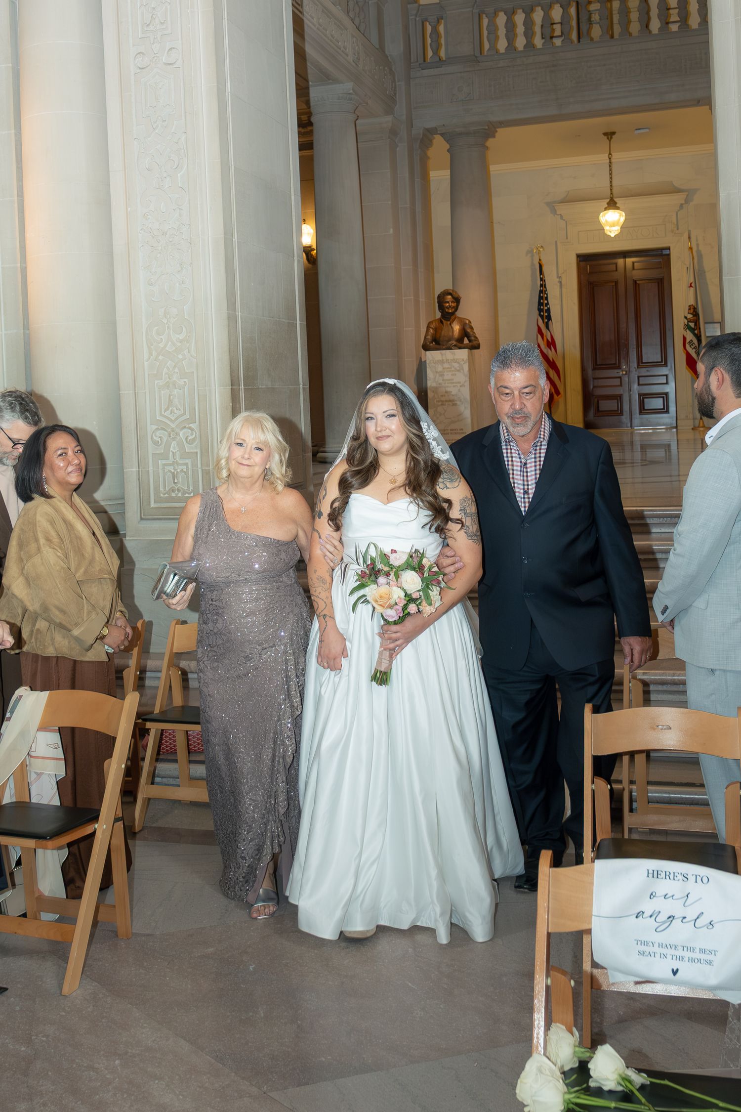Bride walks down the aisle in white gown with bouquet, escorted by parents in elegant wedding ceremony.