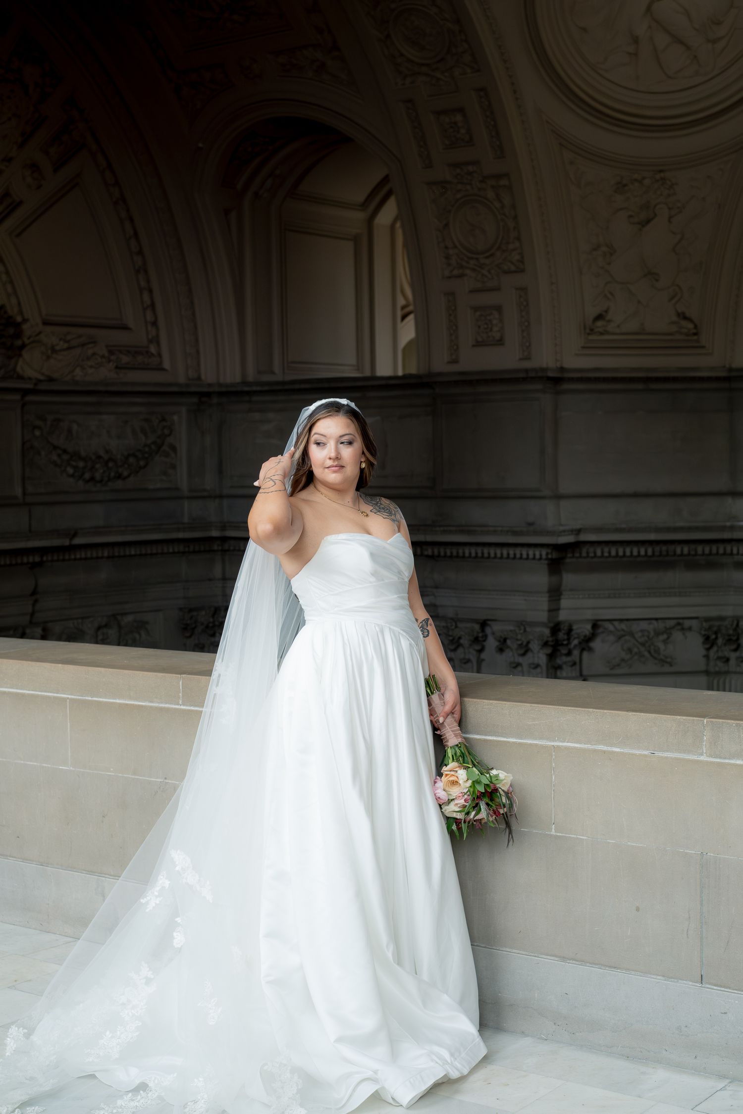 Bride in strapless white gown holding bouquet with flowing veil in ornate building.