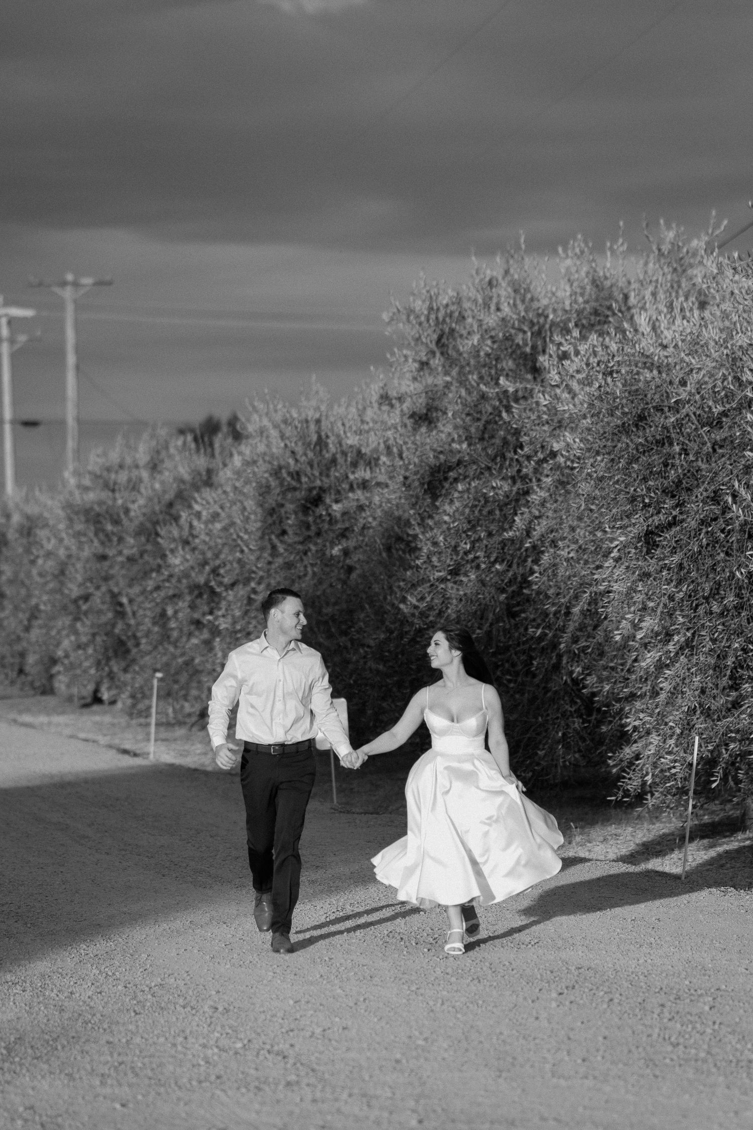 Black and white photo of bride and groom joyfully running hand in hand along a tree-lined gravel path.