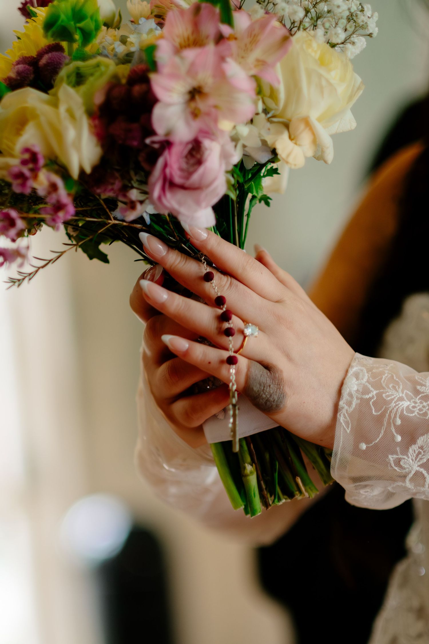 Bride holding a colorful bouquet with a diamond ring and rosary bracelet on her manicured hands.