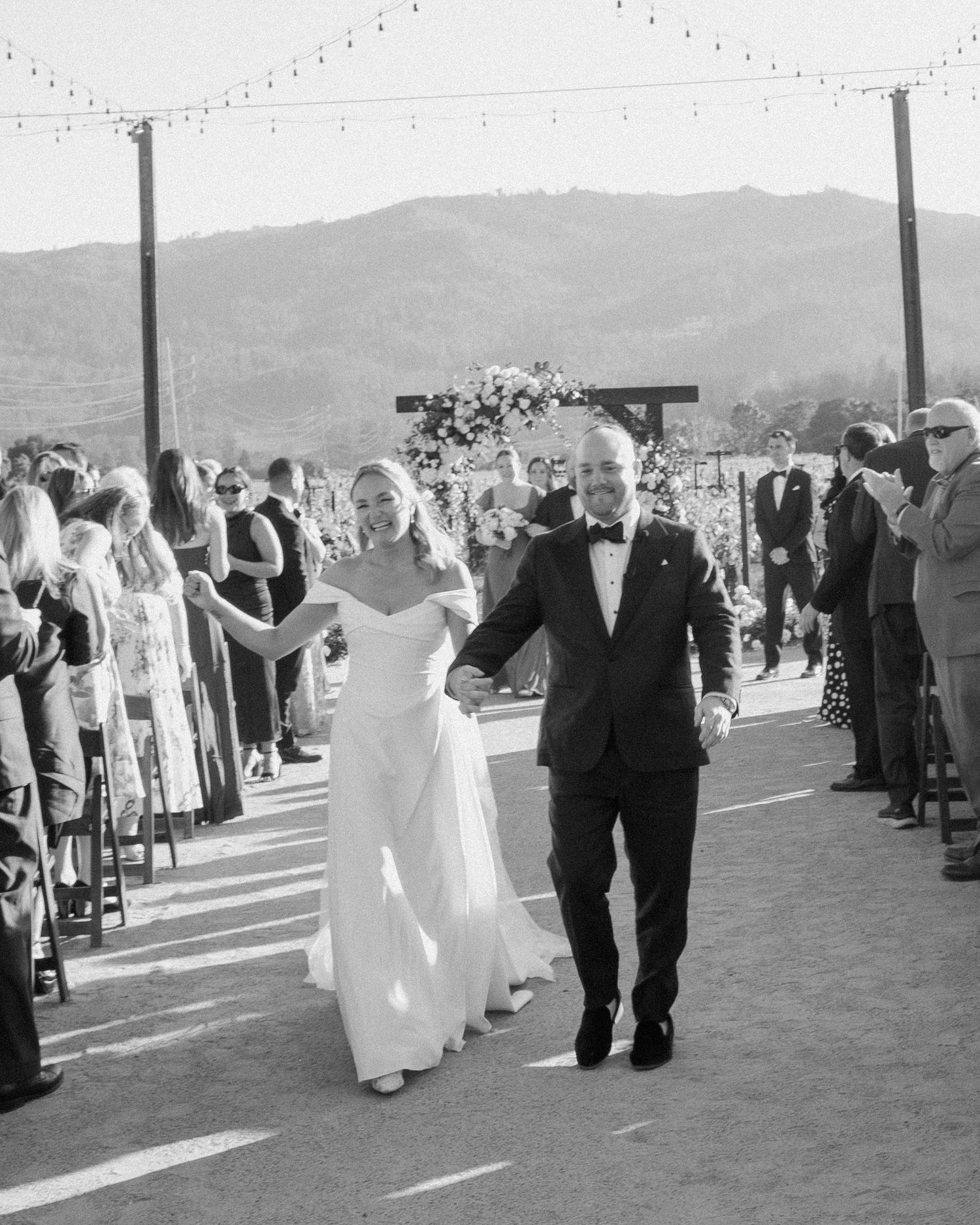 Black and white photo of newlyweds walking down the aisle at an outdoor vineyard wedding ceremony.