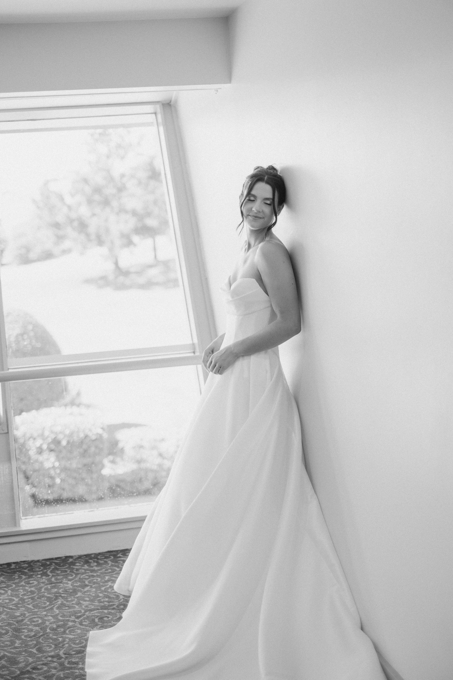 Elegant bride in a strapless ball gown leans against a wall by a window in a black and white portrait.