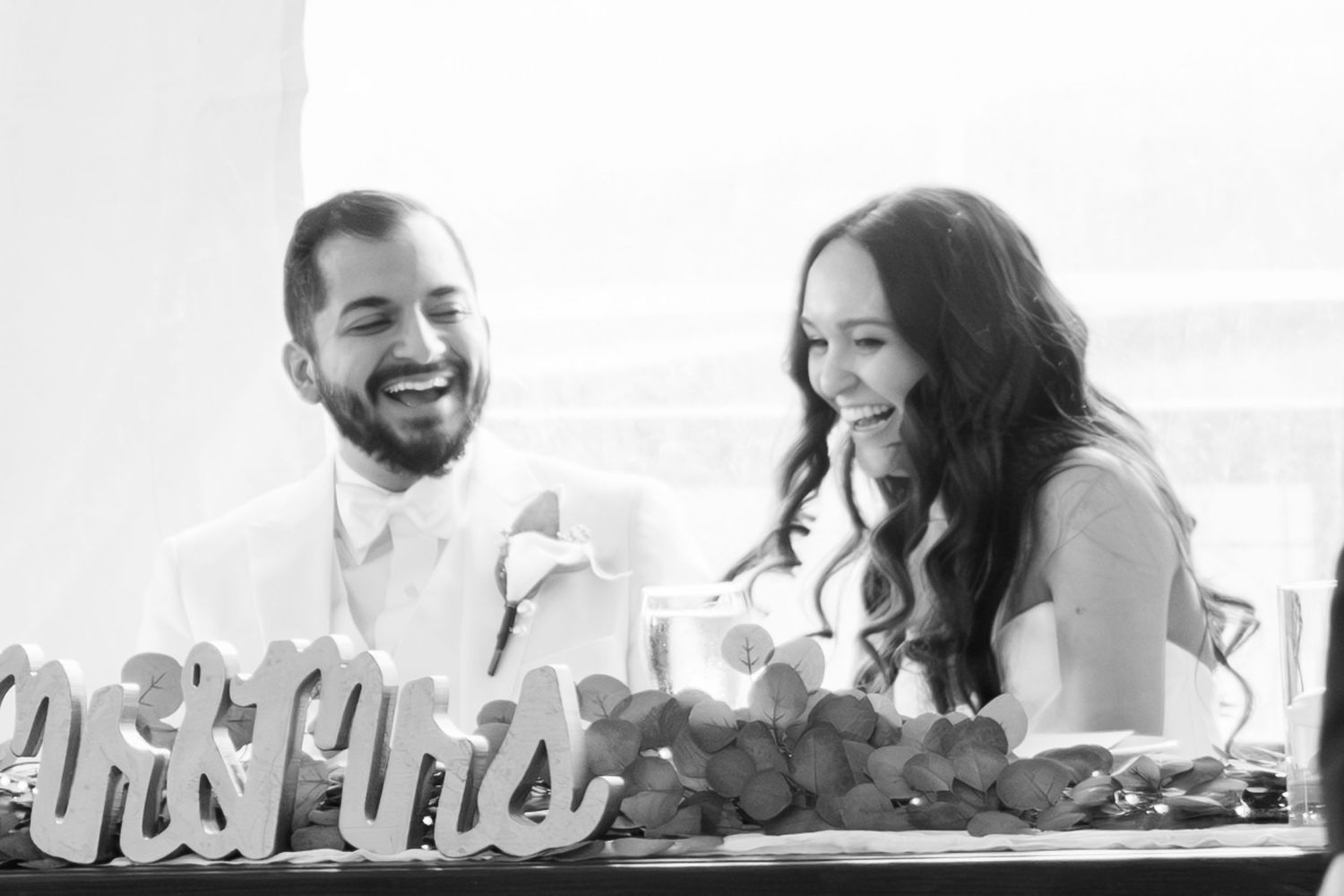 Black and white photo of smiling bride and groom laughing together at sweetheart table with Mr and Mrs sign.
