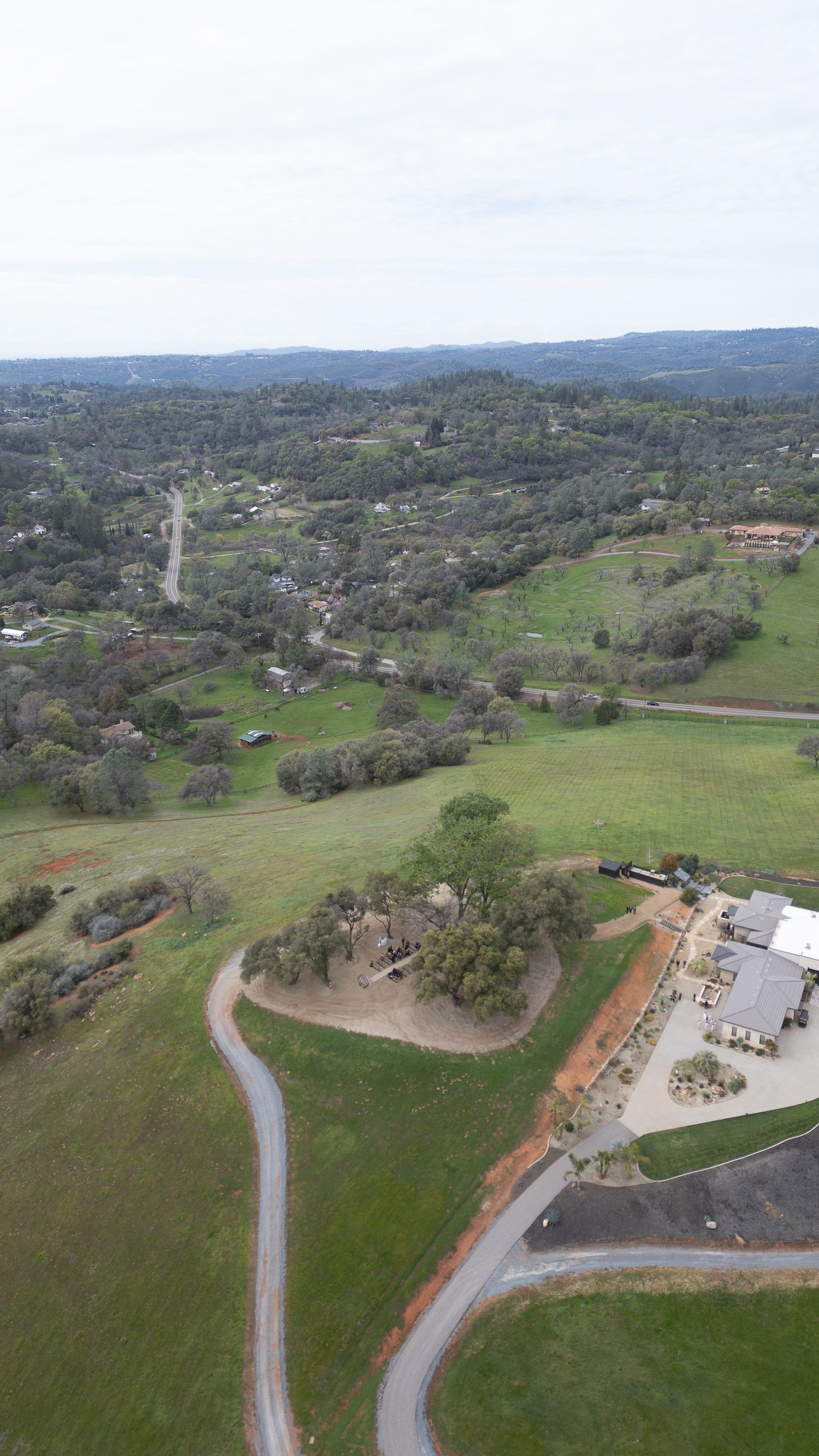 Aerial drone view of a rural valley with winding roads, green fields, scattered trees, and buildings under a cloudy sky.