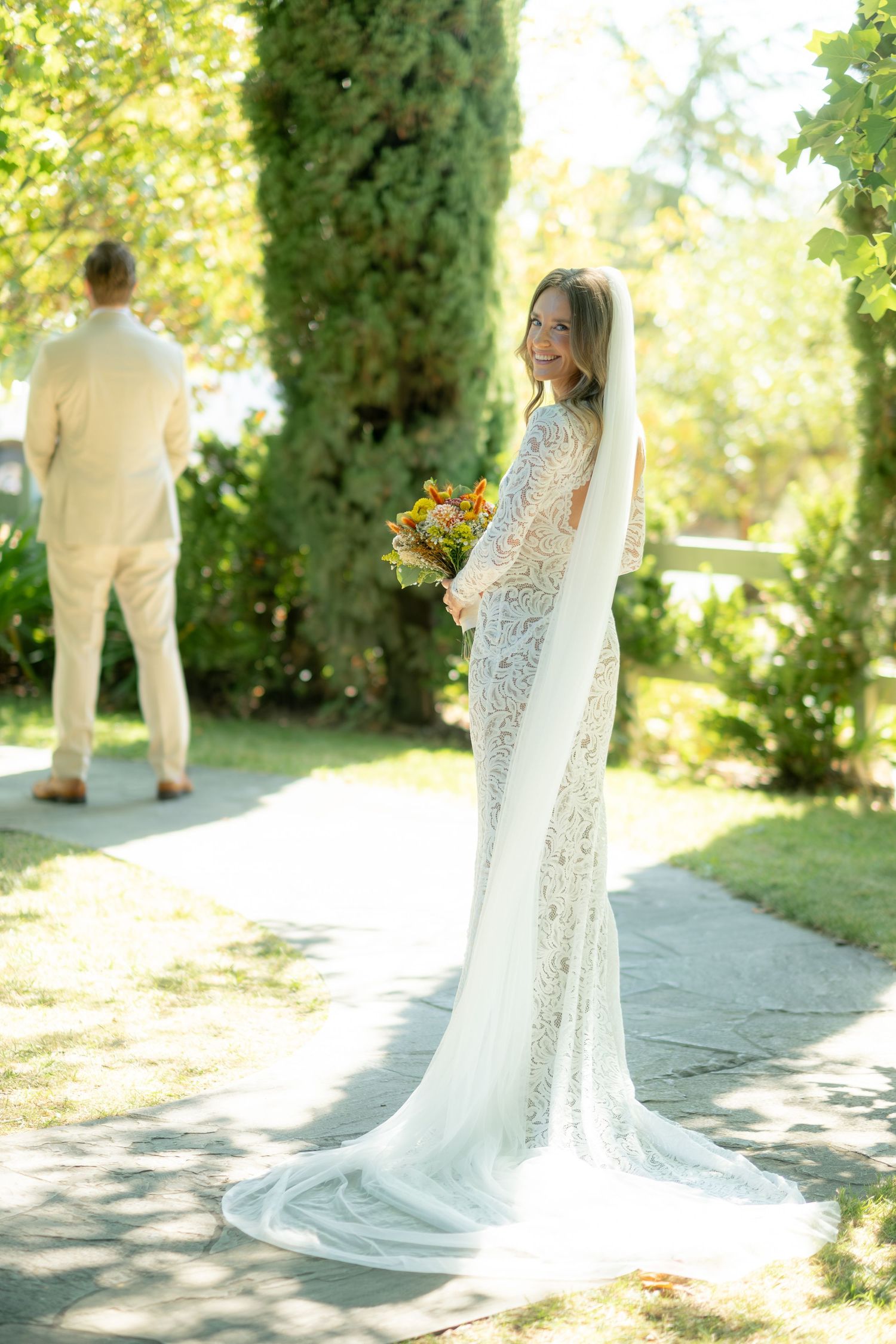 Bride in lace gown with veil smiles over shoulder holding bouquet during first look outdoors.