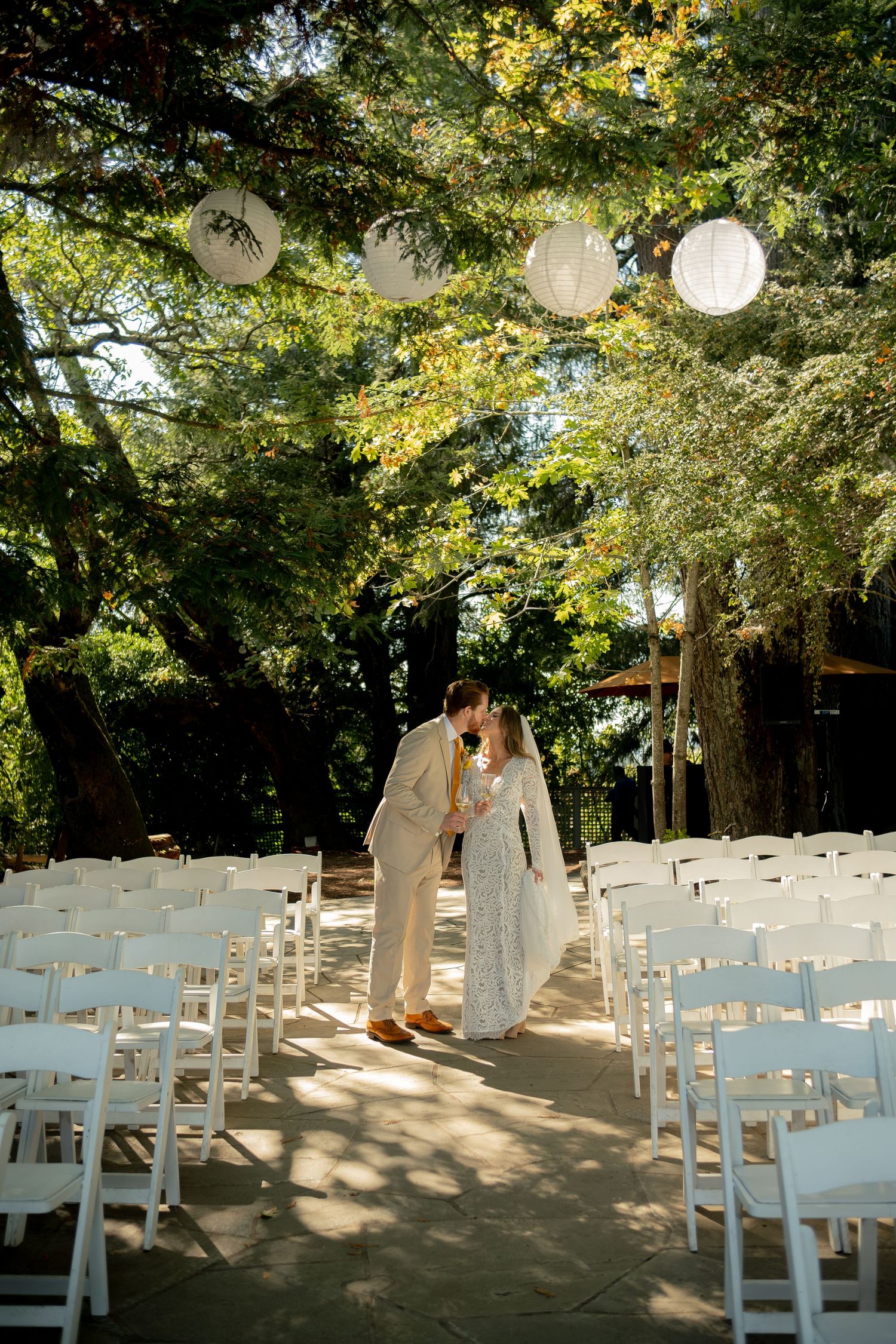 Bride and groom share a kiss at the end of the aisle under white paper lanterns and trees.