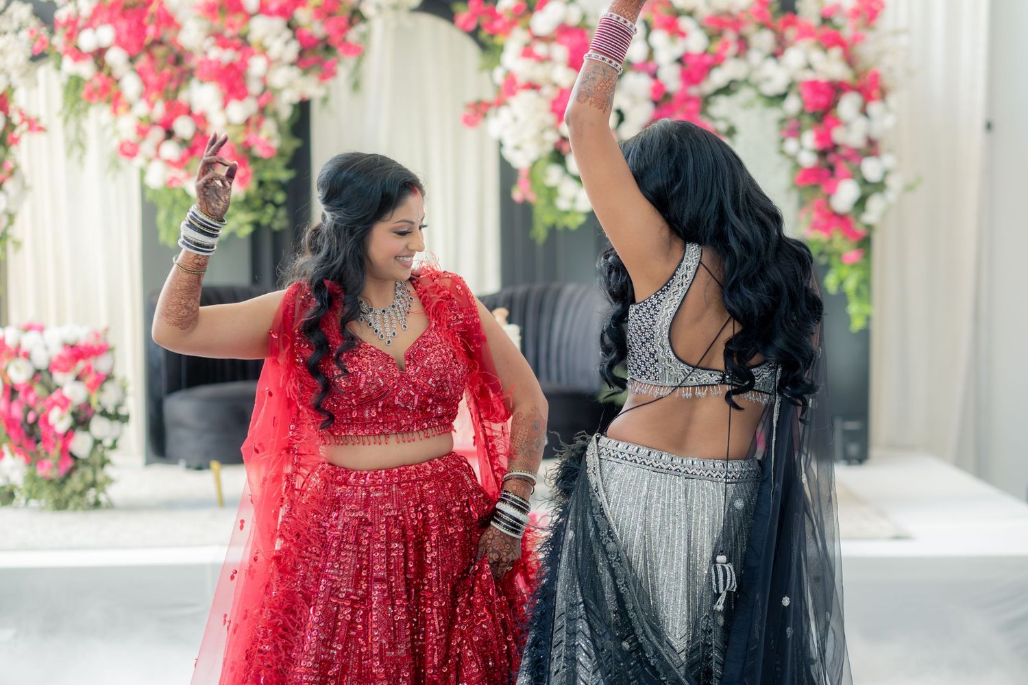 Two women in red and grey lehengas dancing at an Indian wedding celebration.