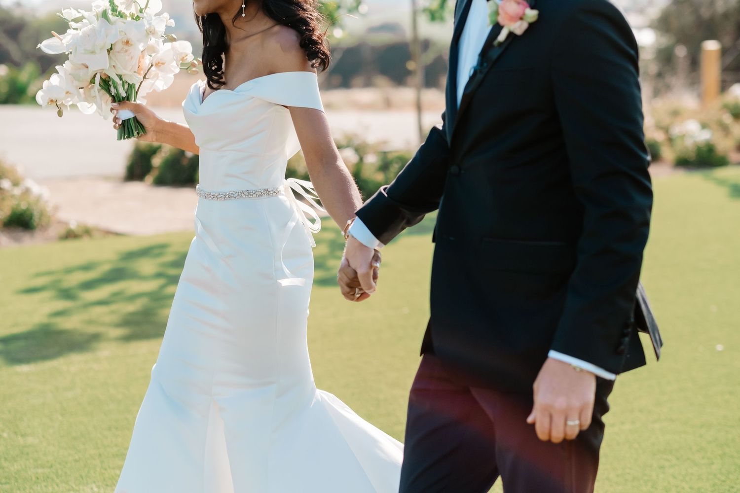 Bride in off-shoulder white gown holding white bouquet walks hand-in-hand with groom in black suit.