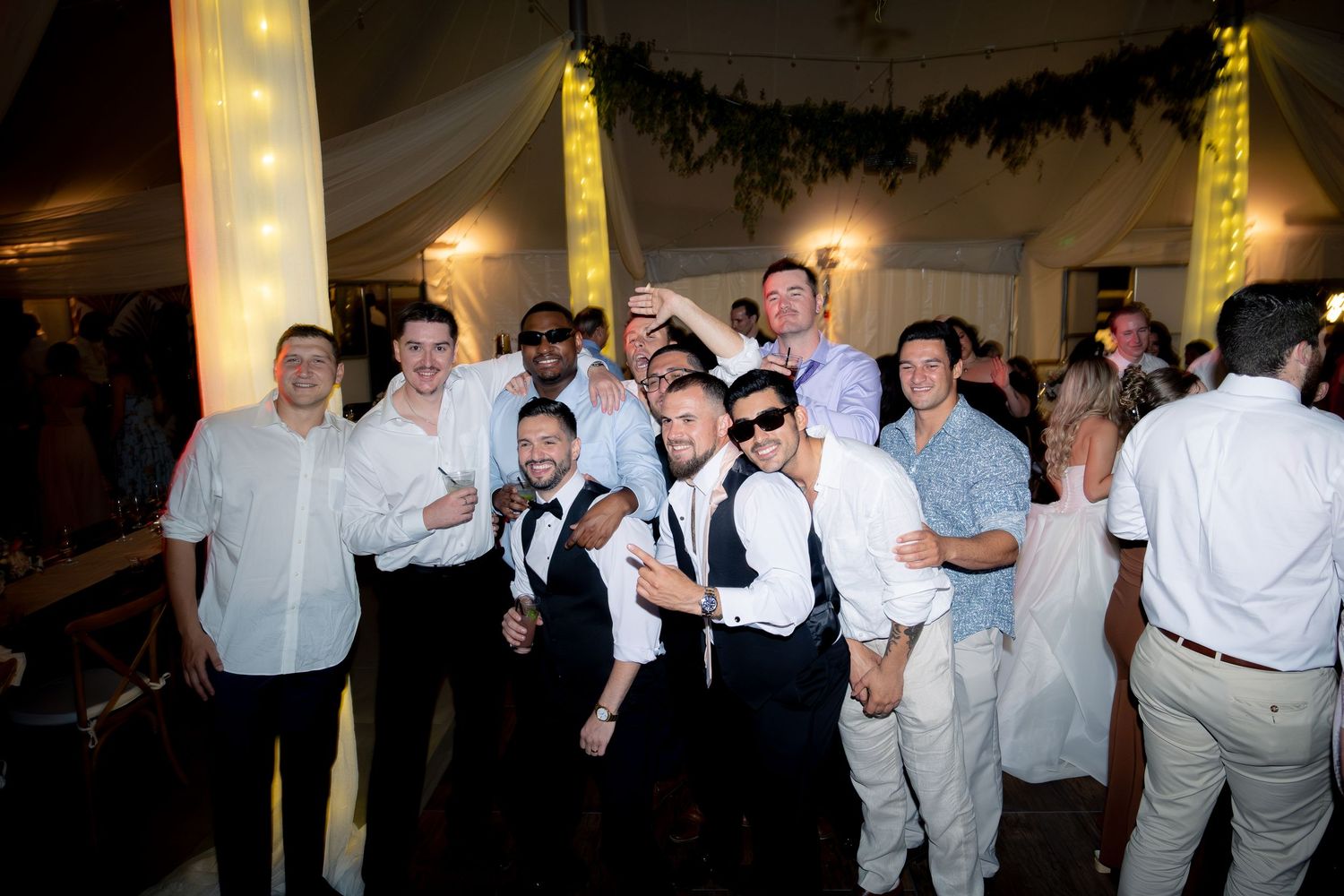 Groom poses with groomsmen and friends for a group photo at elegant wedding reception tent.
