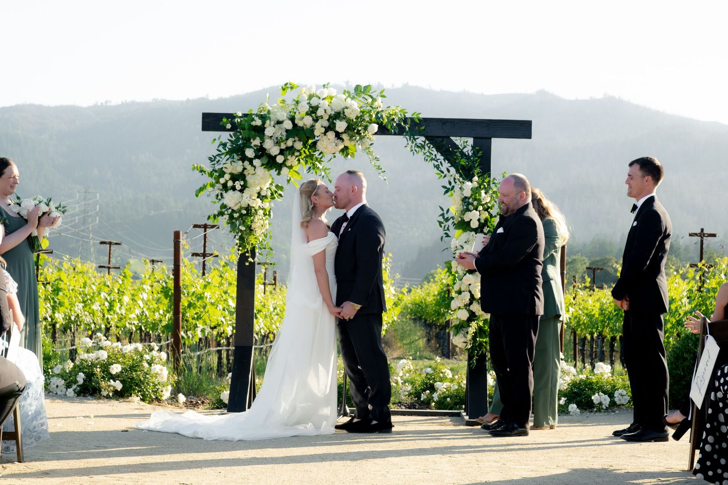 Bride and groom share first kiss under floral arch at outdoor vineyard wedding ceremony.