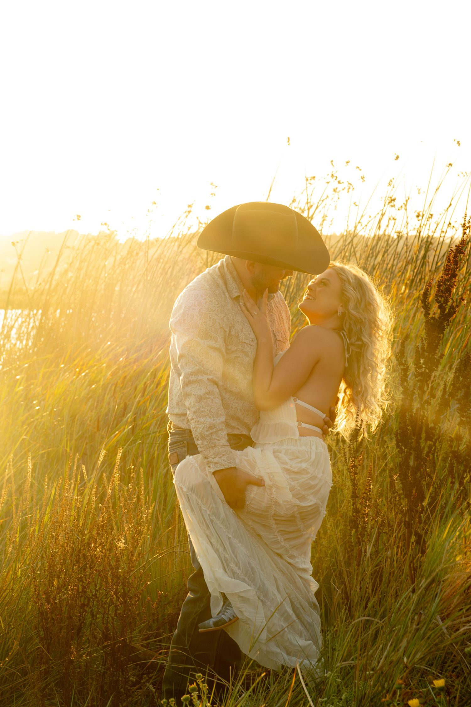 Couple sharing a romantic moment in a golden sunlit field, man in cowboy hat lifting woman in lace dress.