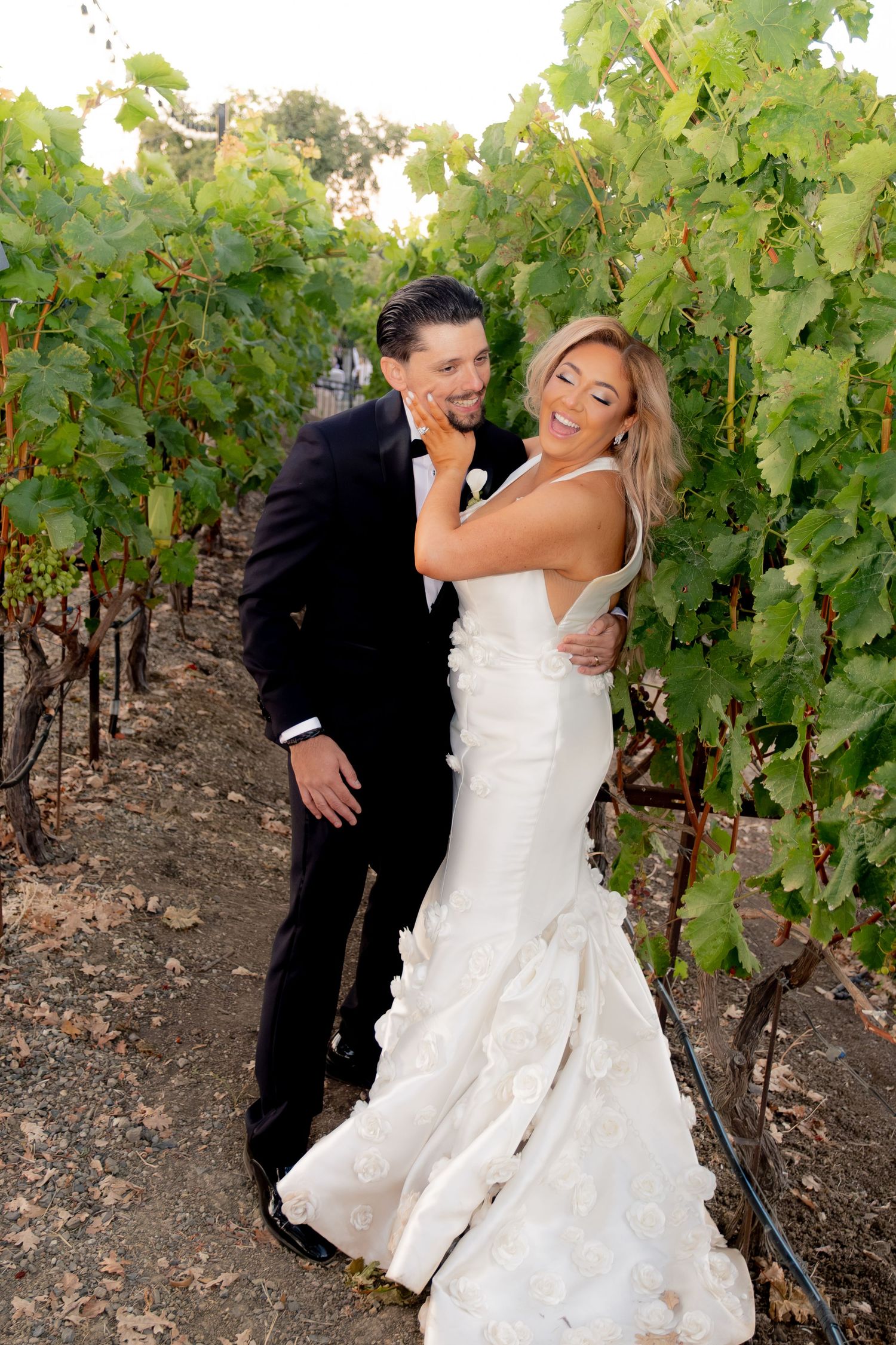 Bride and groom embrace romantically among lush green vineyard rows on their wedding day.