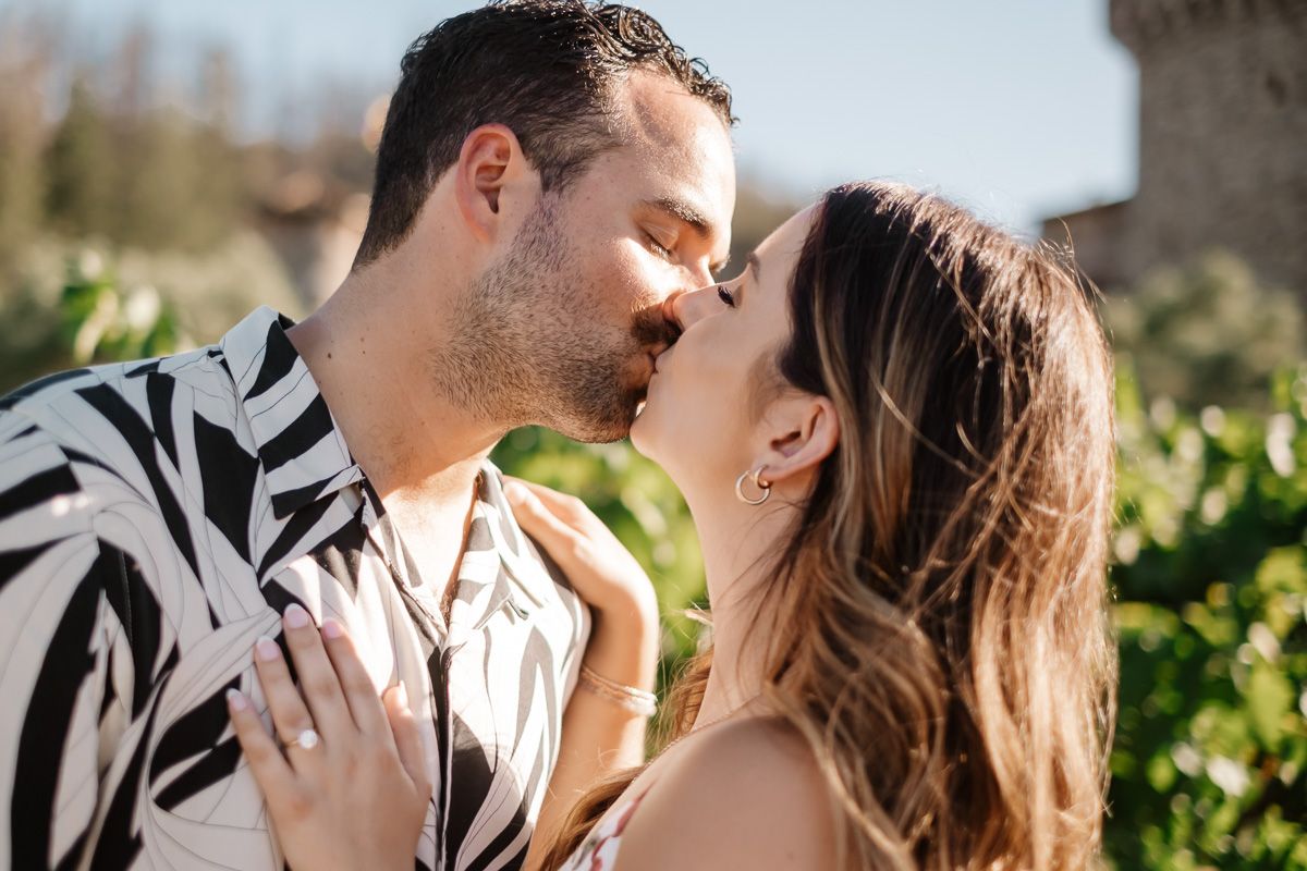 A couple shares a romantic kiss outdoors, with the man in a zebra-print shirt and woman in a beige top.