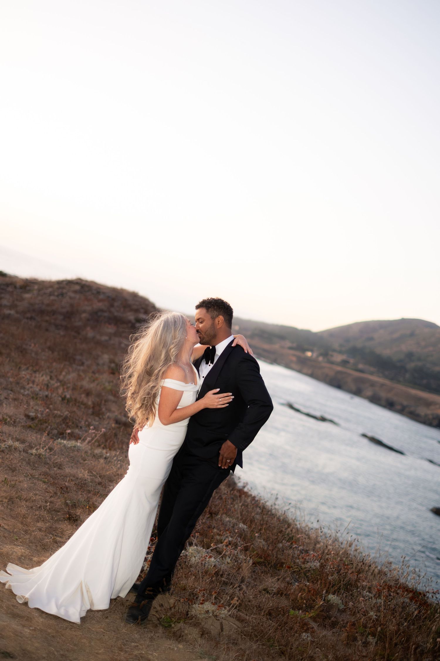 A bride and groom share a romantic kiss on a hillside overlooking a river at sunset.