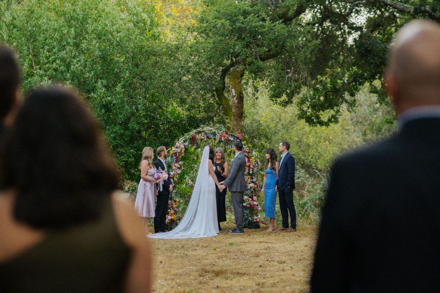 Outdoor wedding ceremony with floral arch, bride and groom, and guests watching in a natural garden setting.