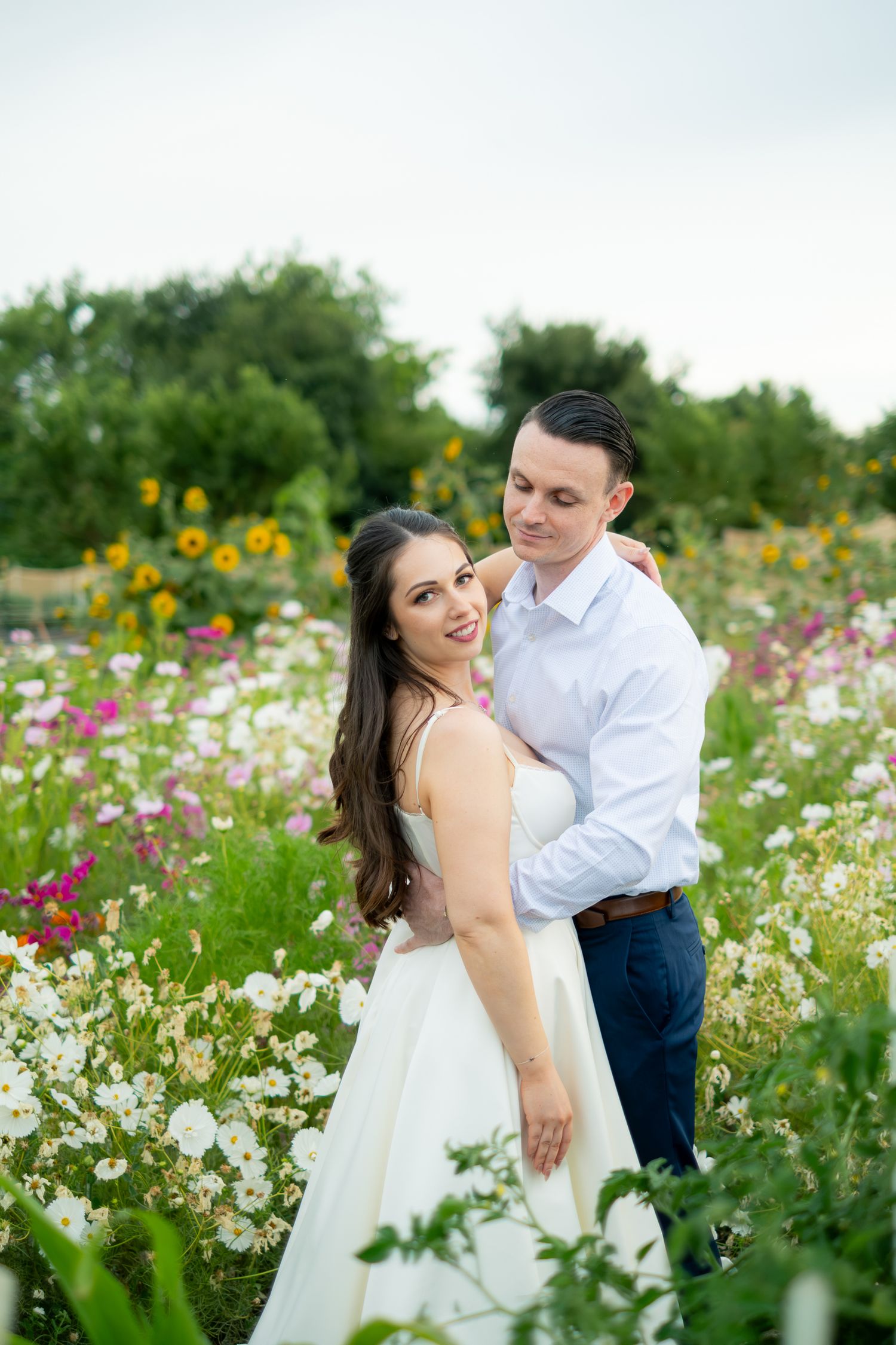 Couple embracing in a colorful wildflower field, bride in white dress smiling at camera.