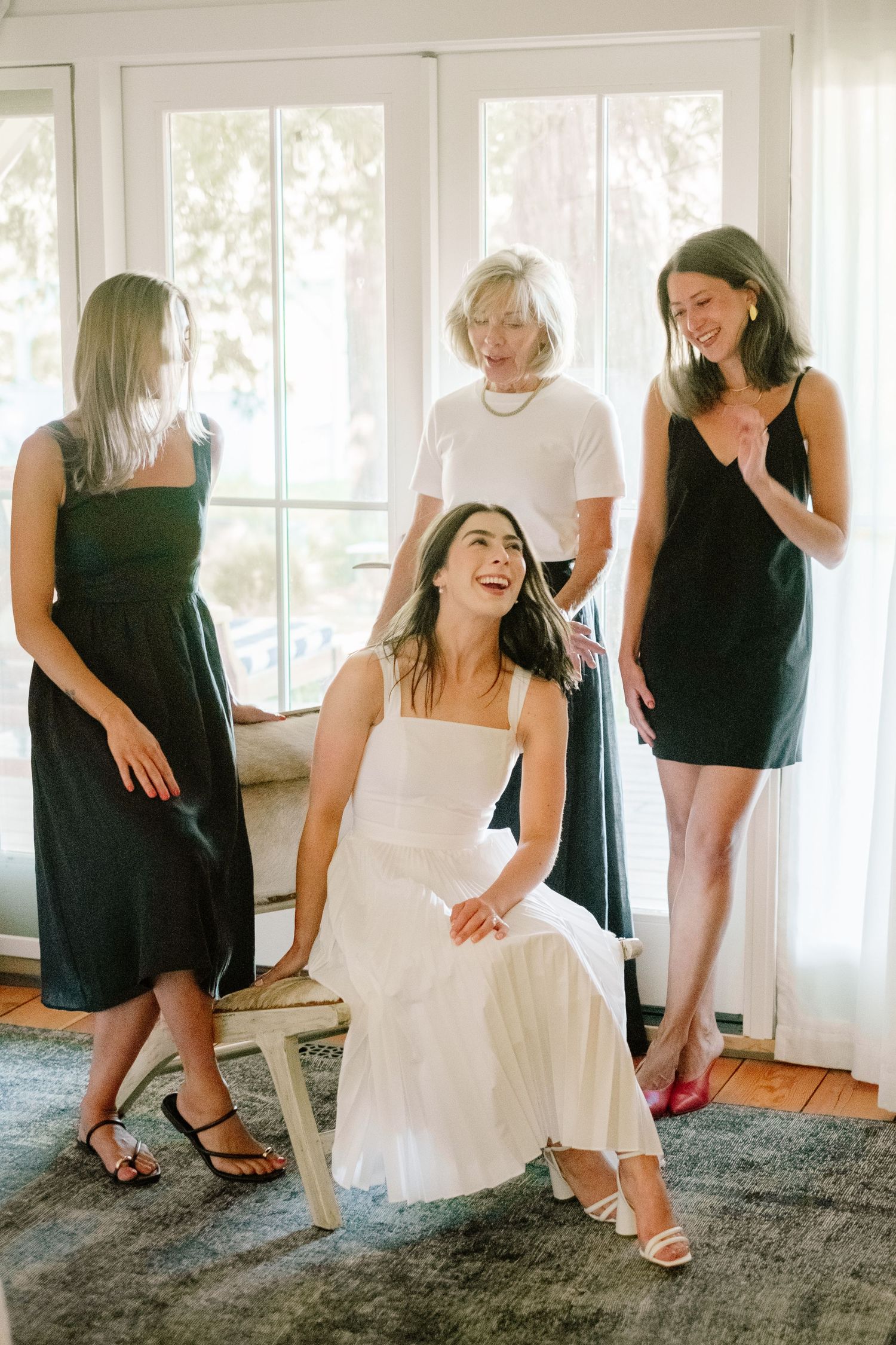 Bride in white dress sits laughing with three women in black dresses during wedding preparations indoors.