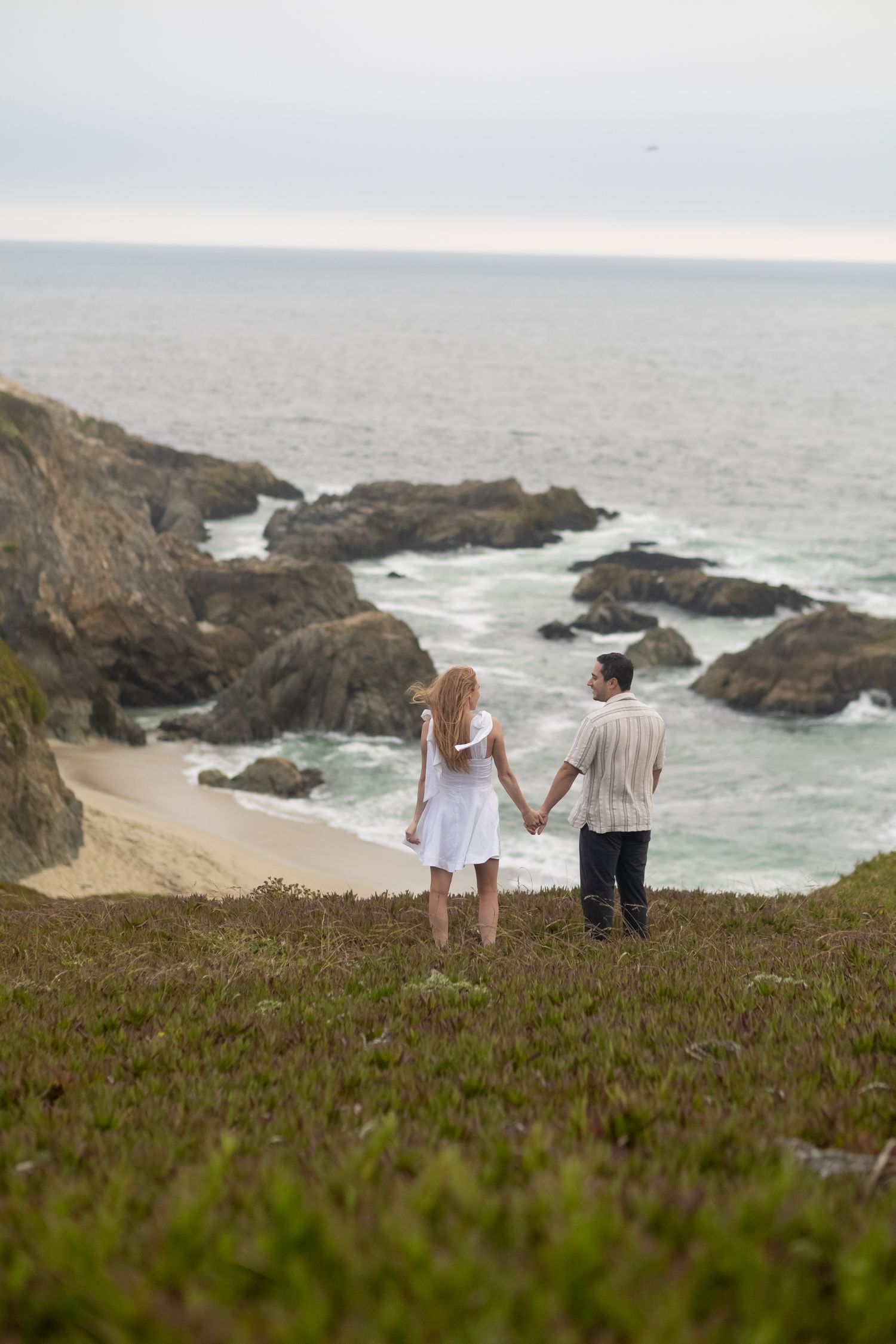 Couple holding hands overlooking a rocky coastal beach with ocean waves from a hillside.