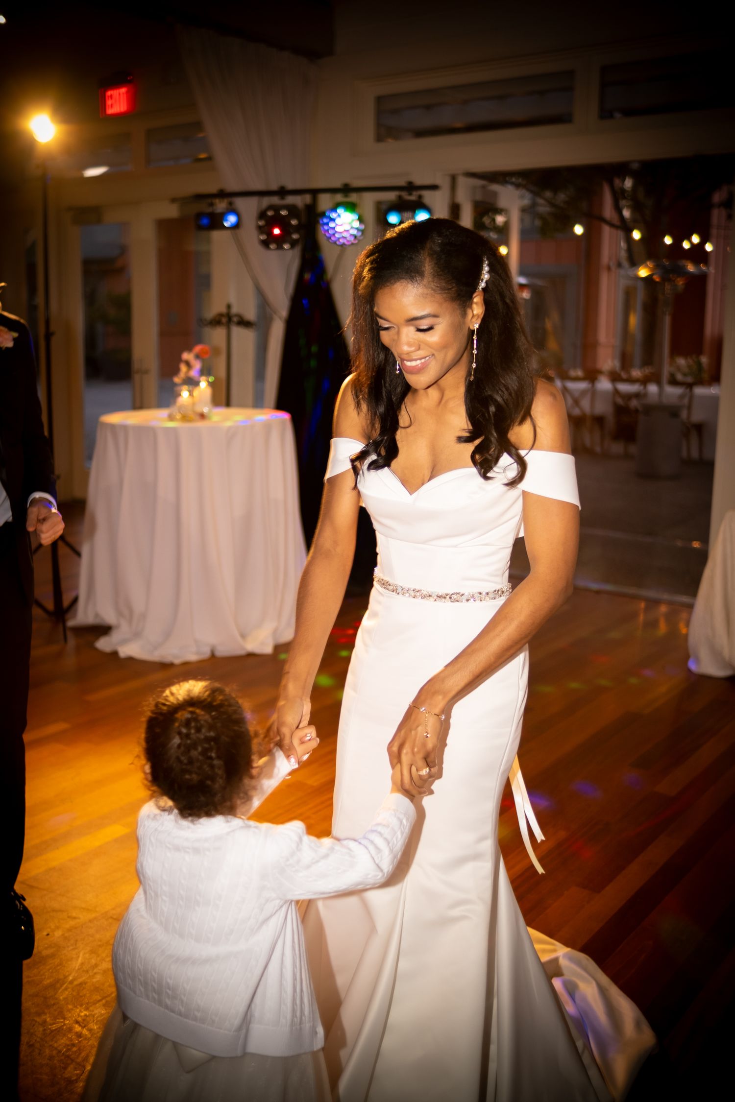 Bride in off-shoulder white gown dancing with young child at wedding reception.