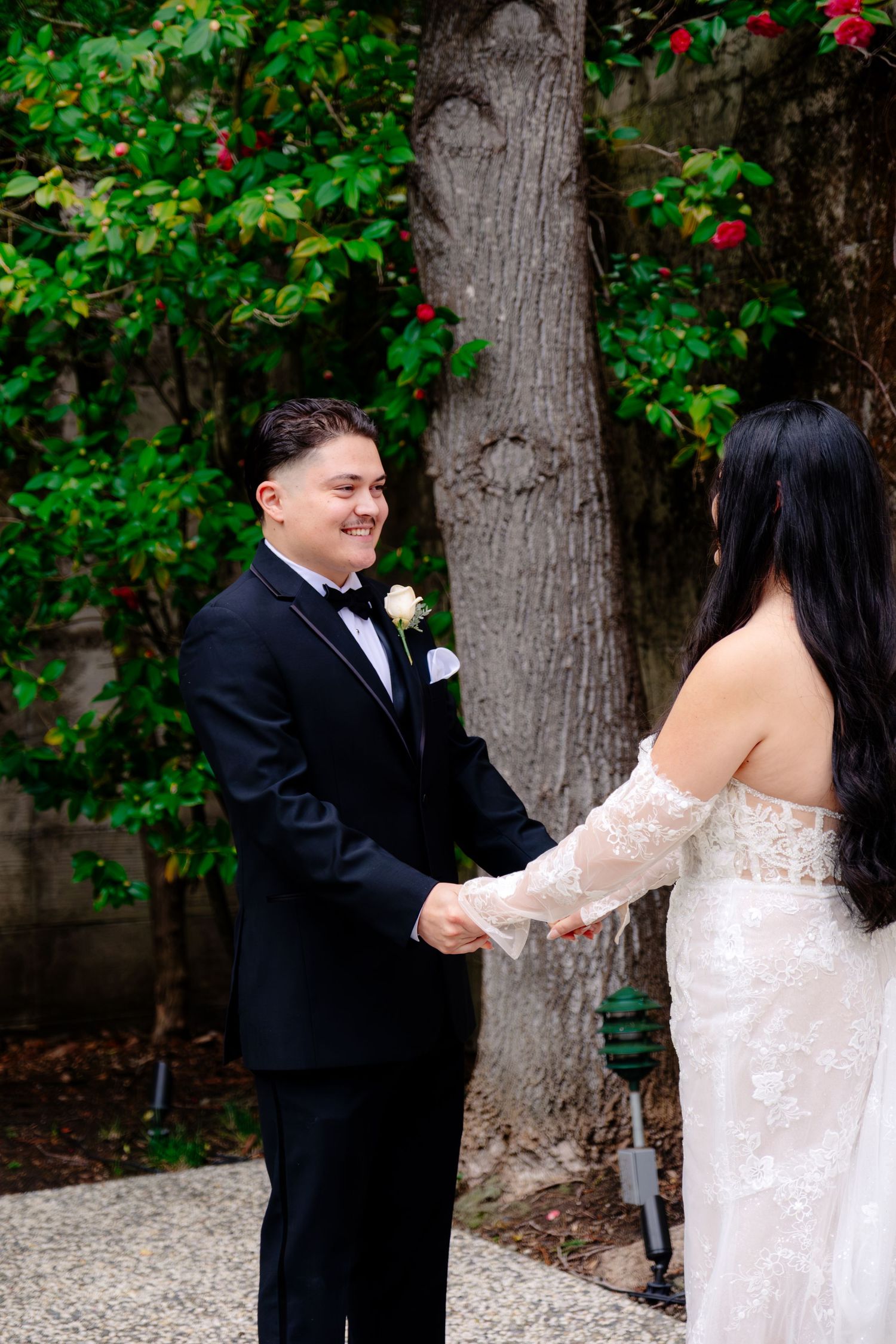 Groom in black tuxedo smiles at bride in lace off-shoulder gown during outdoor first look ceremony.