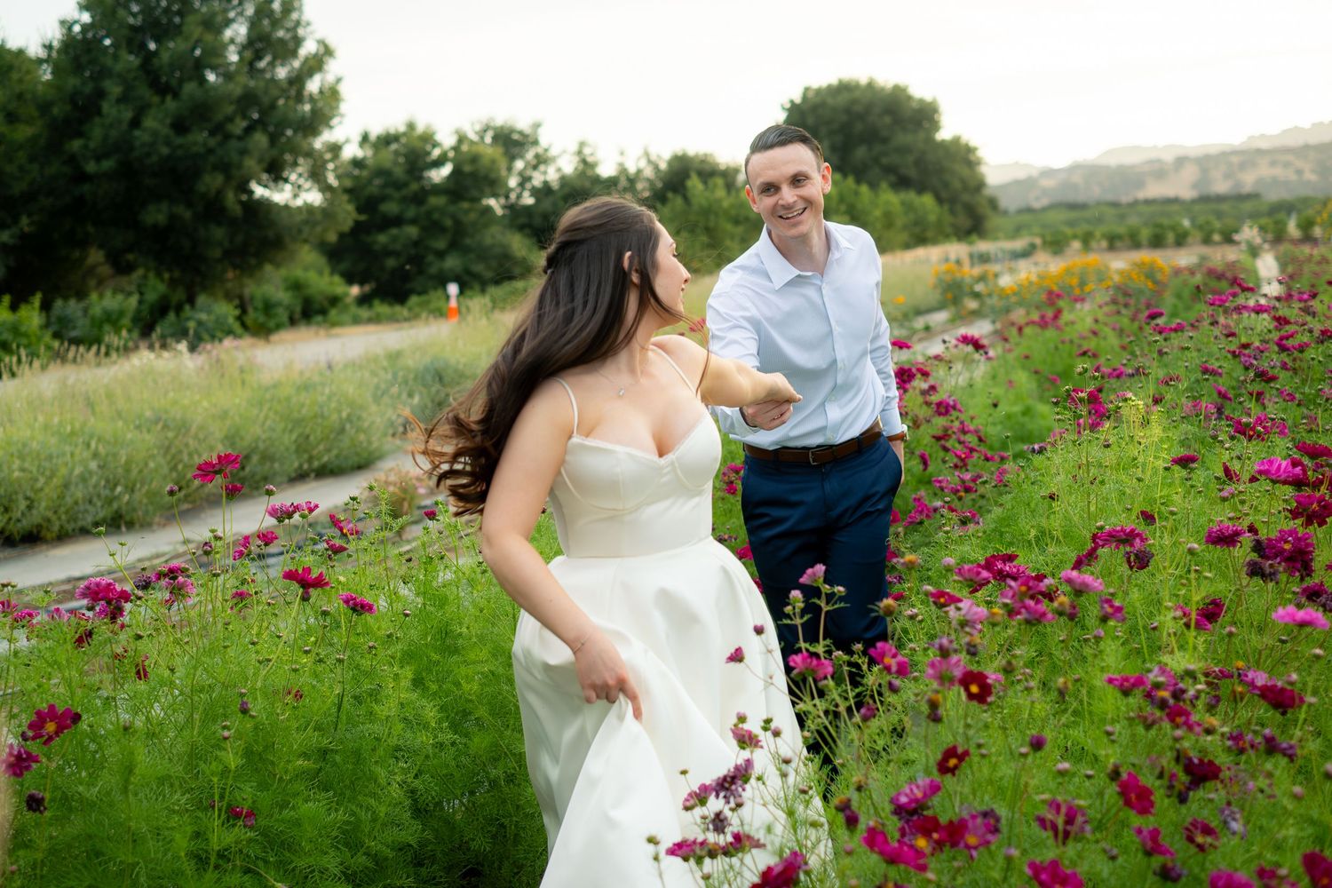 A bride and groom hold hands while walking through a vibrant flower field with pink blooms during their wedding day.