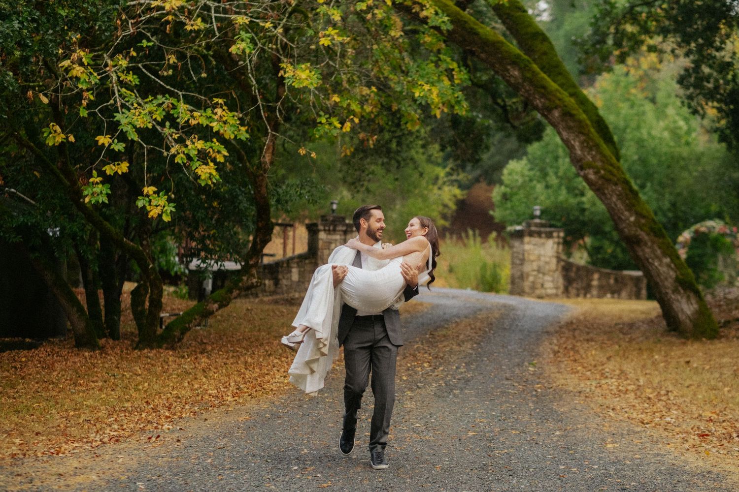 Groom carrying bride in wedding dress on a tree-lined gravel path surrounded by autumn foliage.